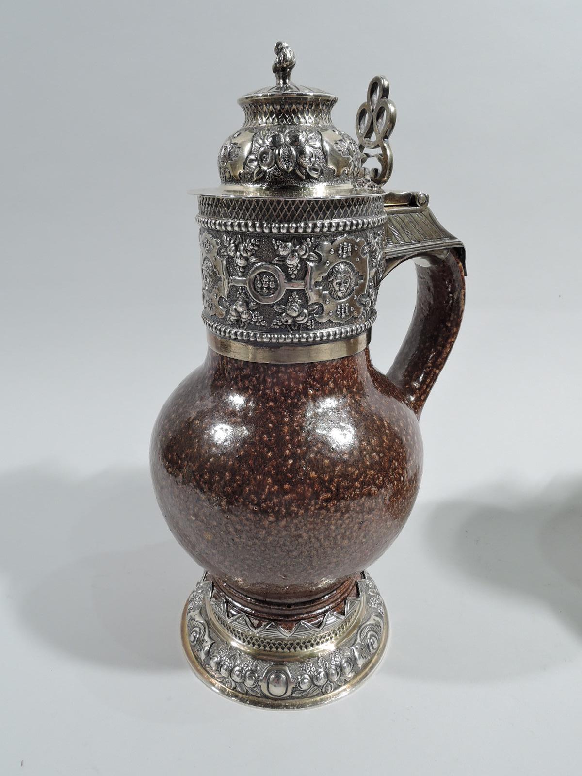 Pair of English Jacobean Revival flagons. Globular body with scroll handle in brown pottery with granulated salt glaze. Gilt sterling silver neck collar and hinged and domed cover with open scrollwork thumb rest as well as raised foot. Chased