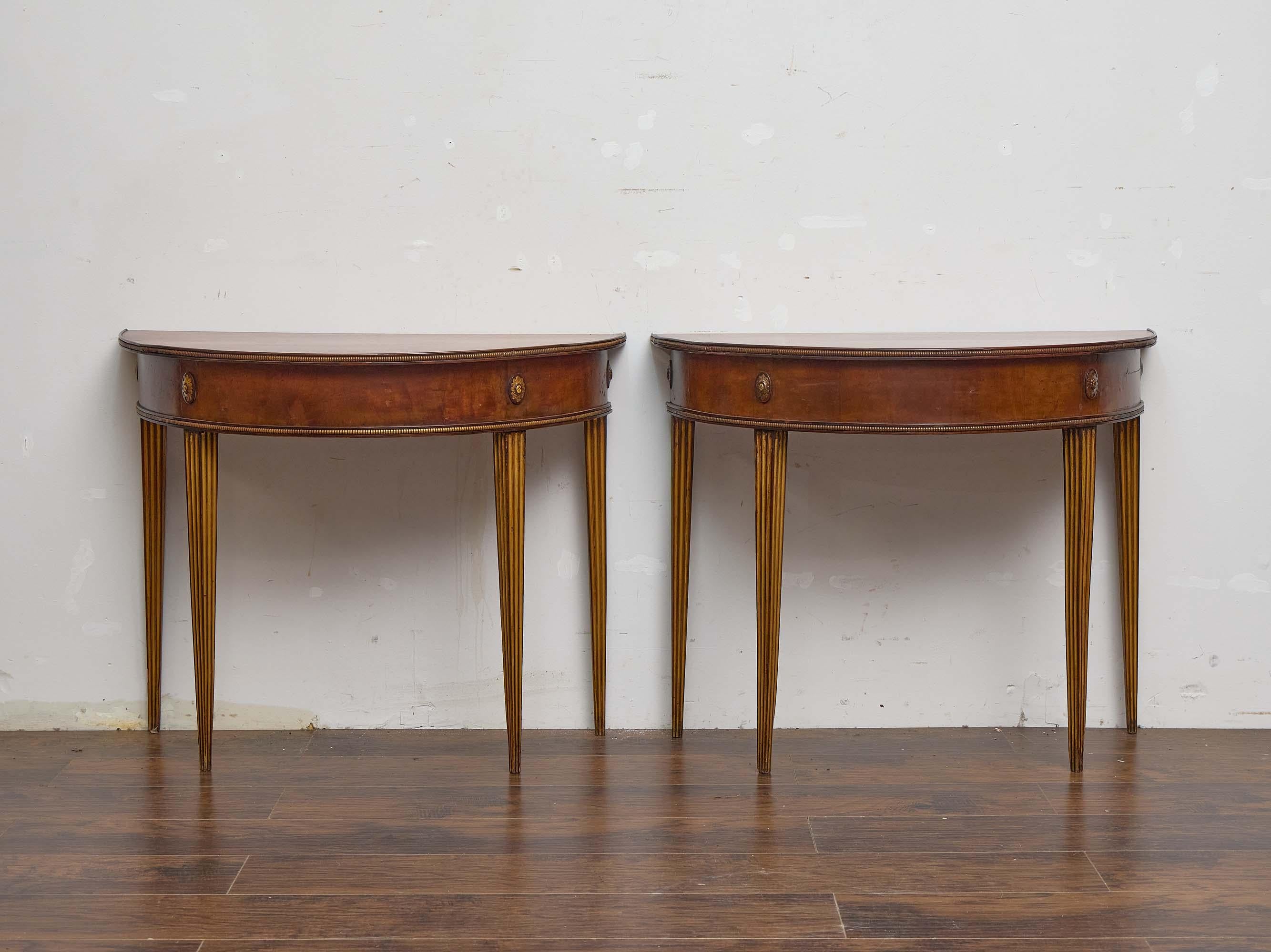 An elegant pair of late Georgian English demi-lune console tables, crafted in mahogany around 1800. Each table features a demi-lune form with a simple veneered top over a shaped apron enriched with carved and painted floral medallions. The aprons