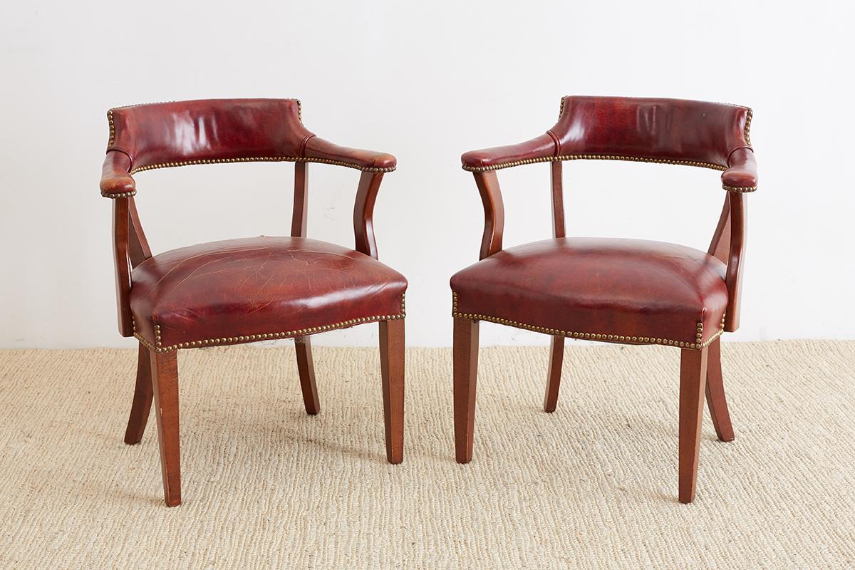 Handsome pair of English mahogany and studded leather captains chairs or studded chairs. Distinctive style finished in dark leather accented by brass nail head trim. Beautiful patina with worn seats and noticeable wear on creases. Also known as bow