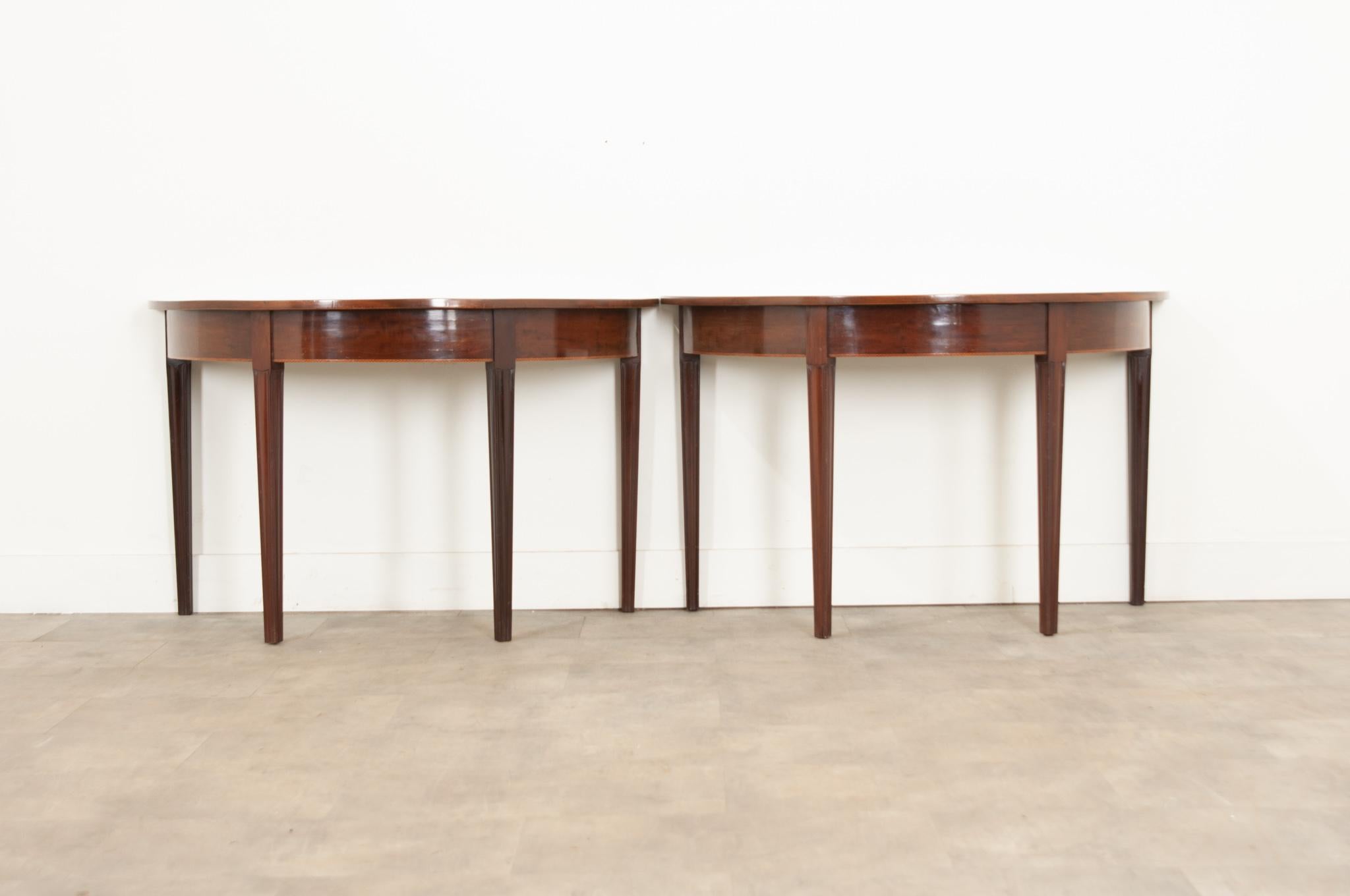 A pair of classic English mahogany demilune console tables with inlay banding and four tapered legs circa 1860. Created in England during the 19th century, each of this pair of demilunes features a semi-circular top sitting above a simple apron made