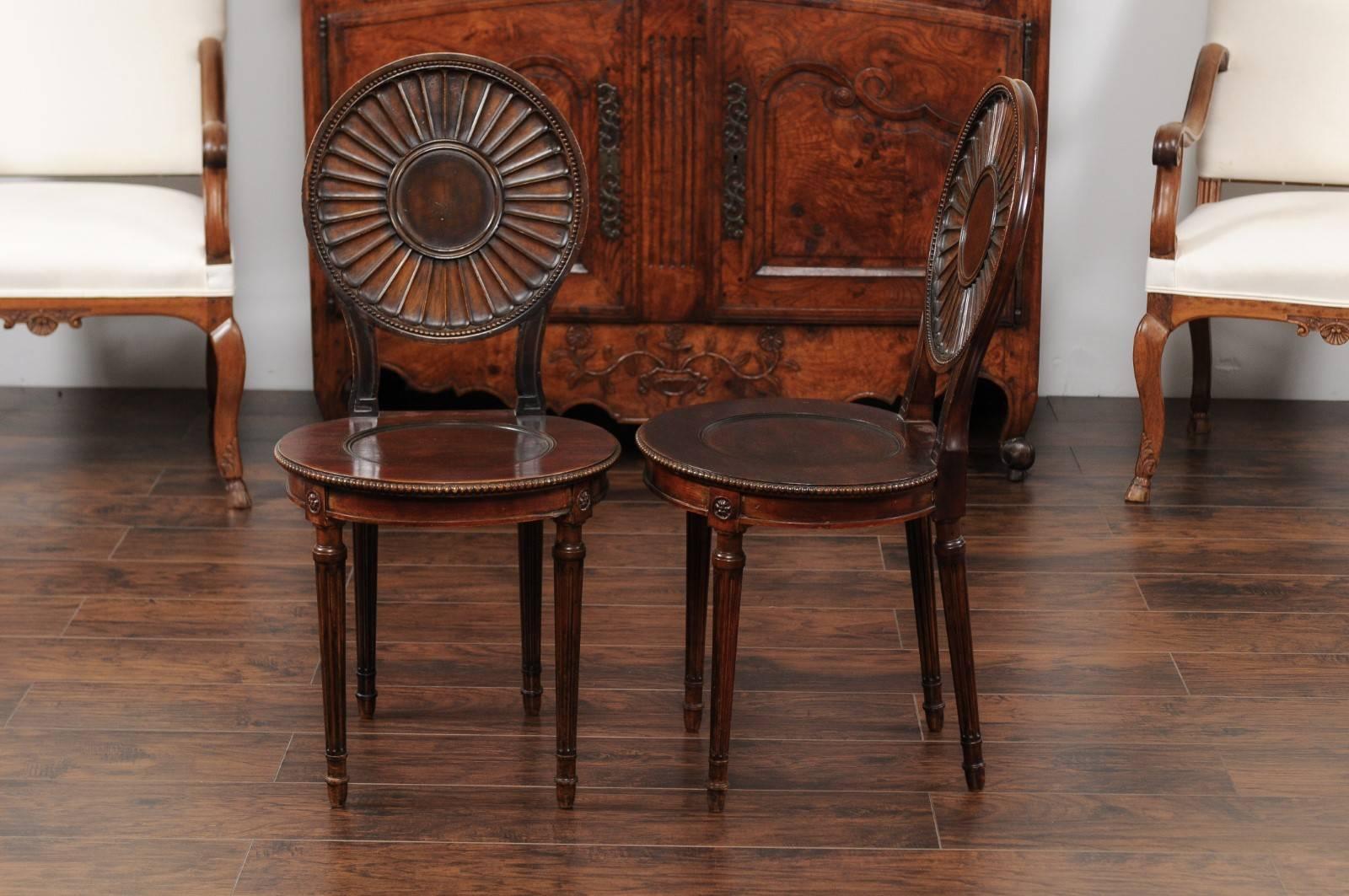 A pair of English mahogany hall chairs from the second half of the 19th century, with round backs and fluted legs. Each of this pair of English hall chairs features an eye-catching slightly slanted round back, adorned with a central medallion from