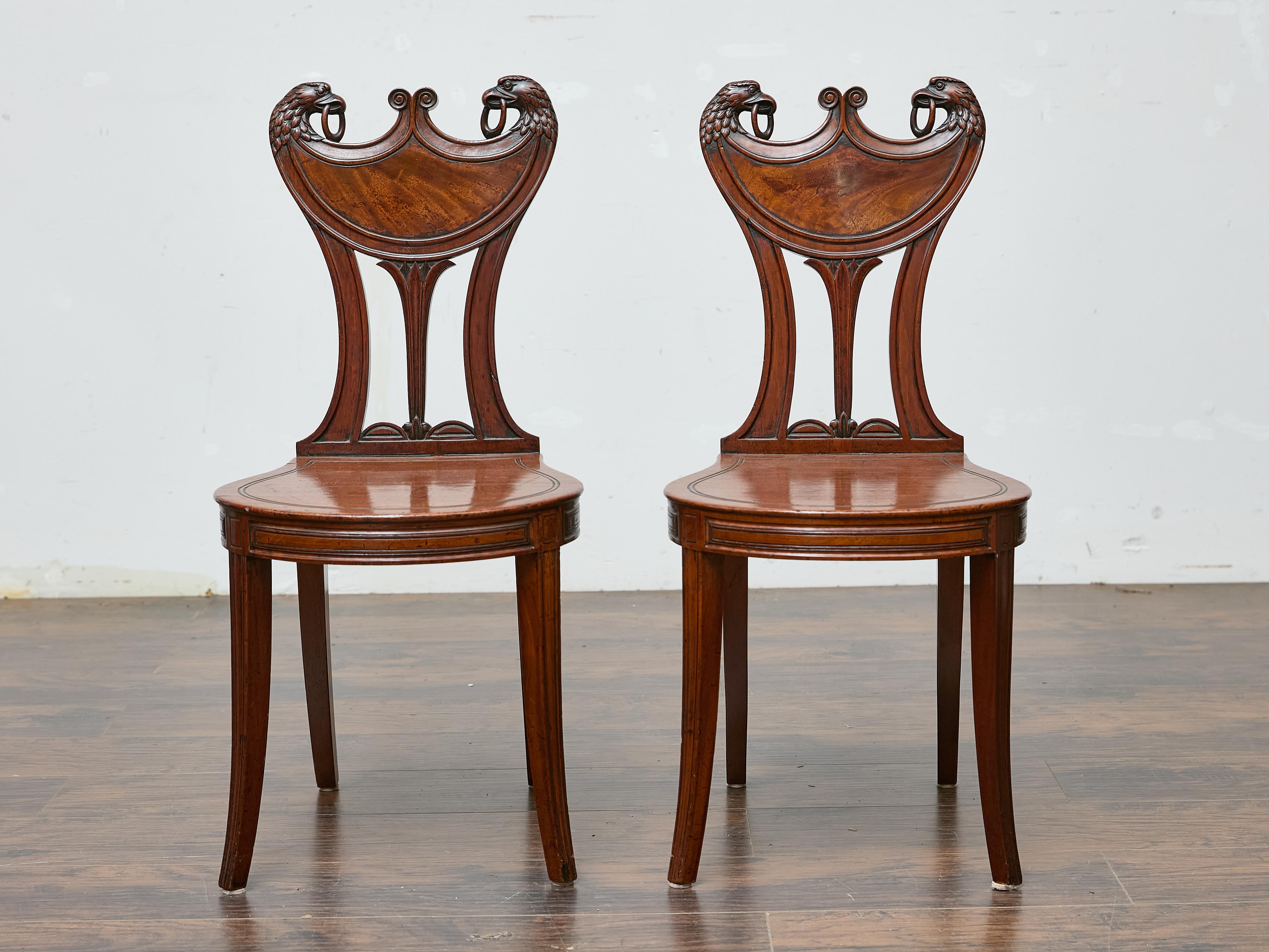 This pair of English mahogany hall chairs dates to the 19th century and stands out for its dramatic carved backrests featuring stylized eagle heads gripping carved rings in their beaks. The back splats, shaped like classical crests, are flanked by