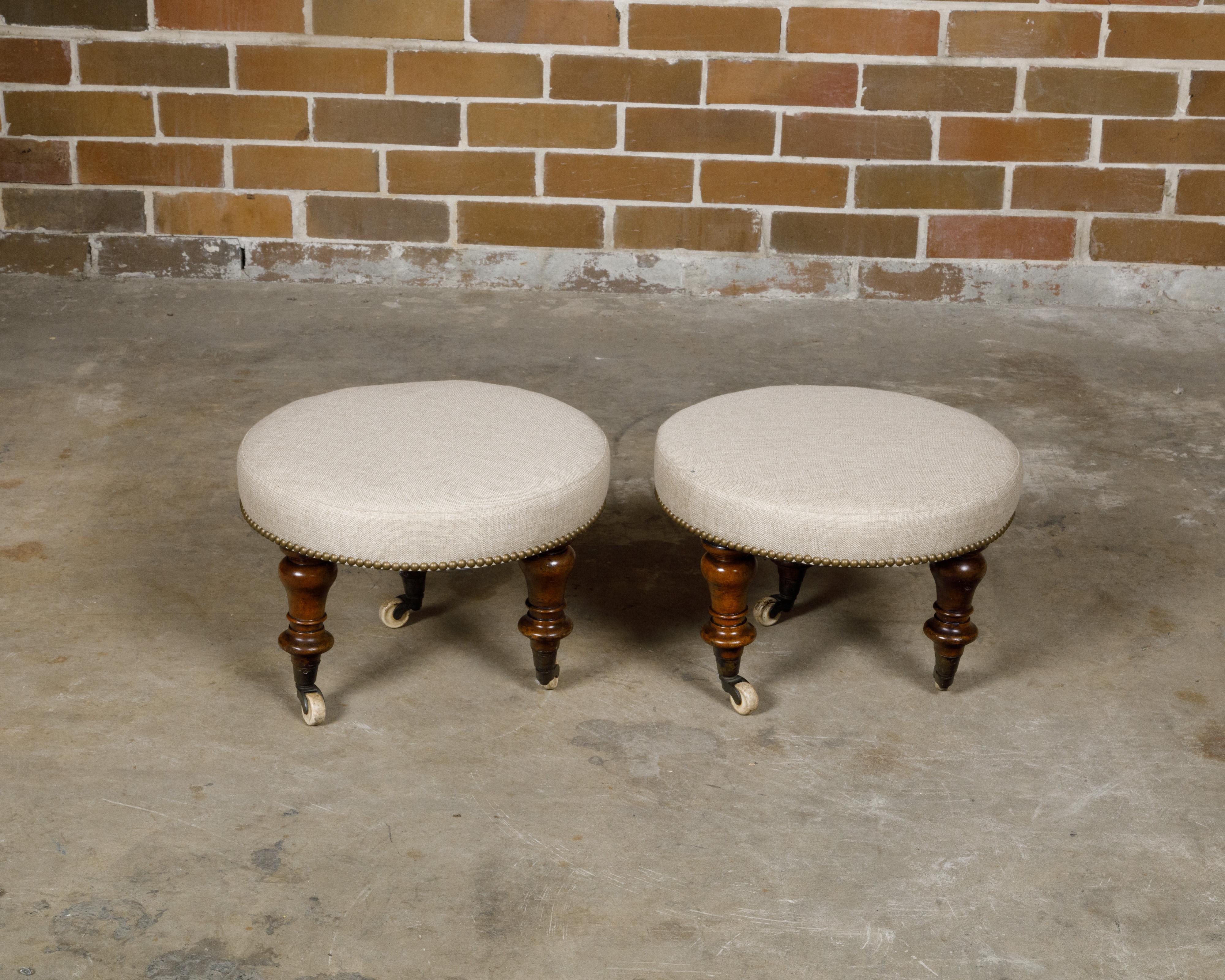 A pair of English mahogany stools from the 19th century with turned legs resting on casters and upholstered seats. This exquisite pair of English mahogany stools features elegantly turned legs sourced from chairs crafted in the 19th century, resting
