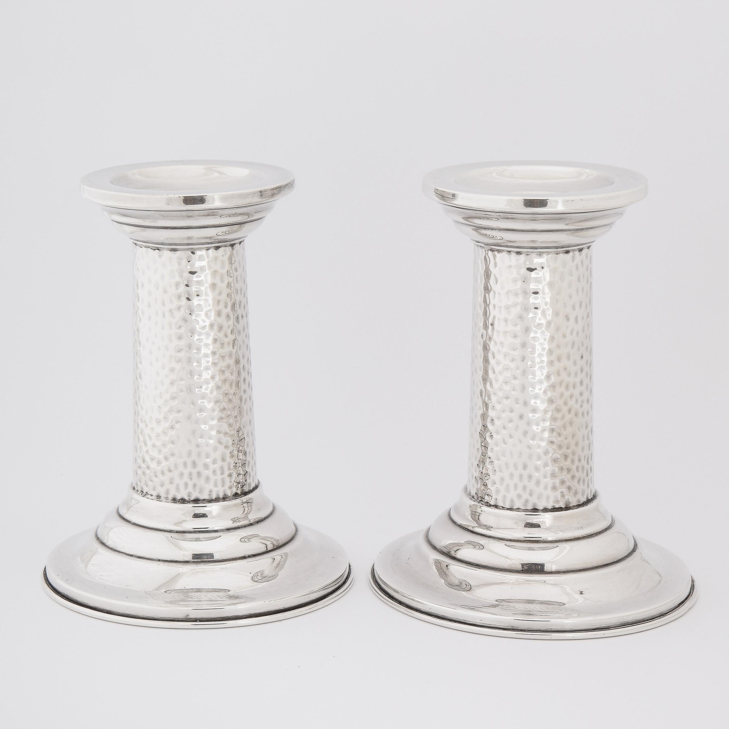 This elegant pair of candlesticks were realized in England circa 1960. They feature tiered concentric circular bases from which hand hammered cylindrical bodies- with a wonderful organic dappled texture- ascend, culminating in circular tiered necks.