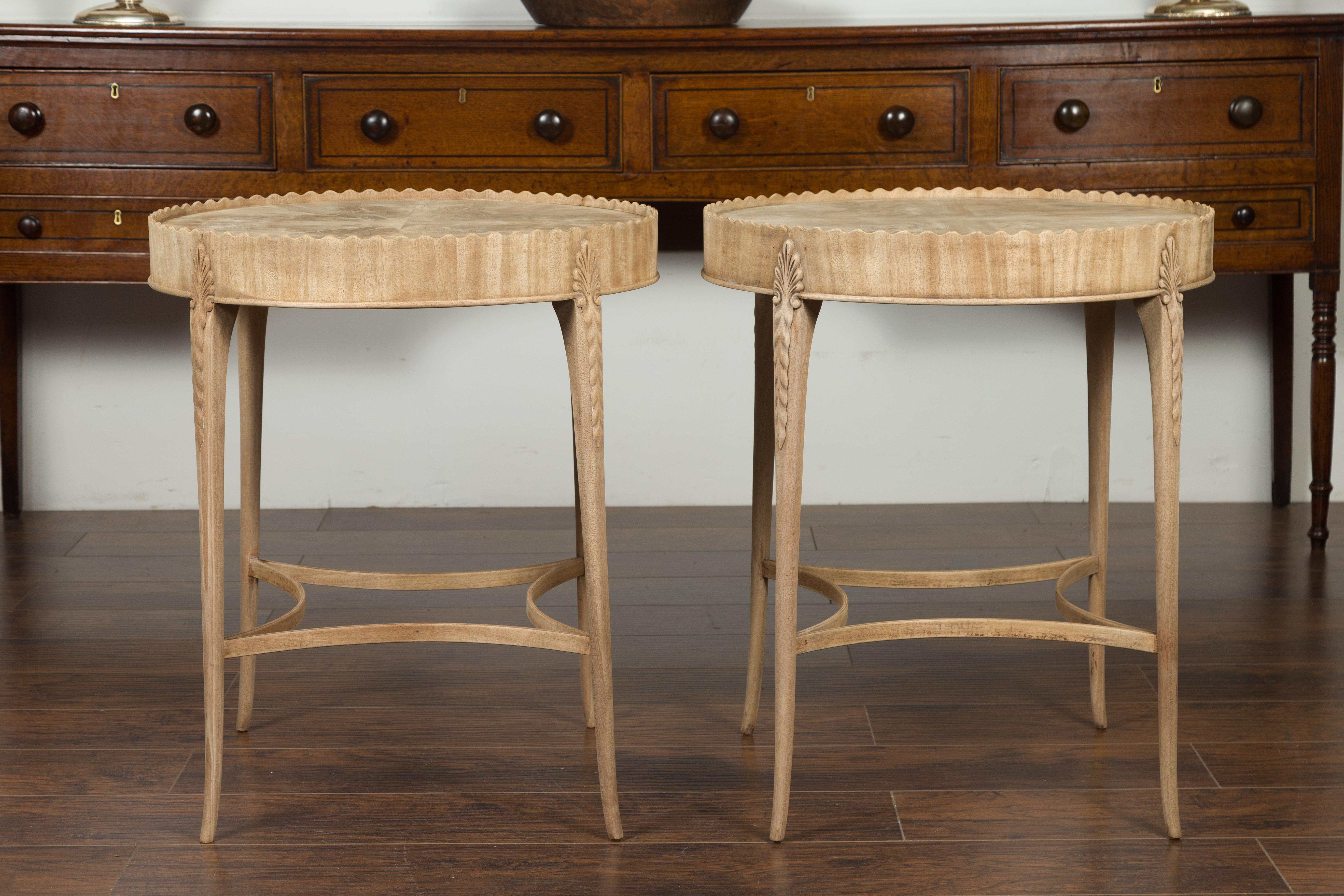 A pair of English vintage bleached mahogany side tables from the mid-20th century, with circular tops, pie crust aprons and carved foliage. Created in England during the midcentury period, each of this pair of side tables features a round top