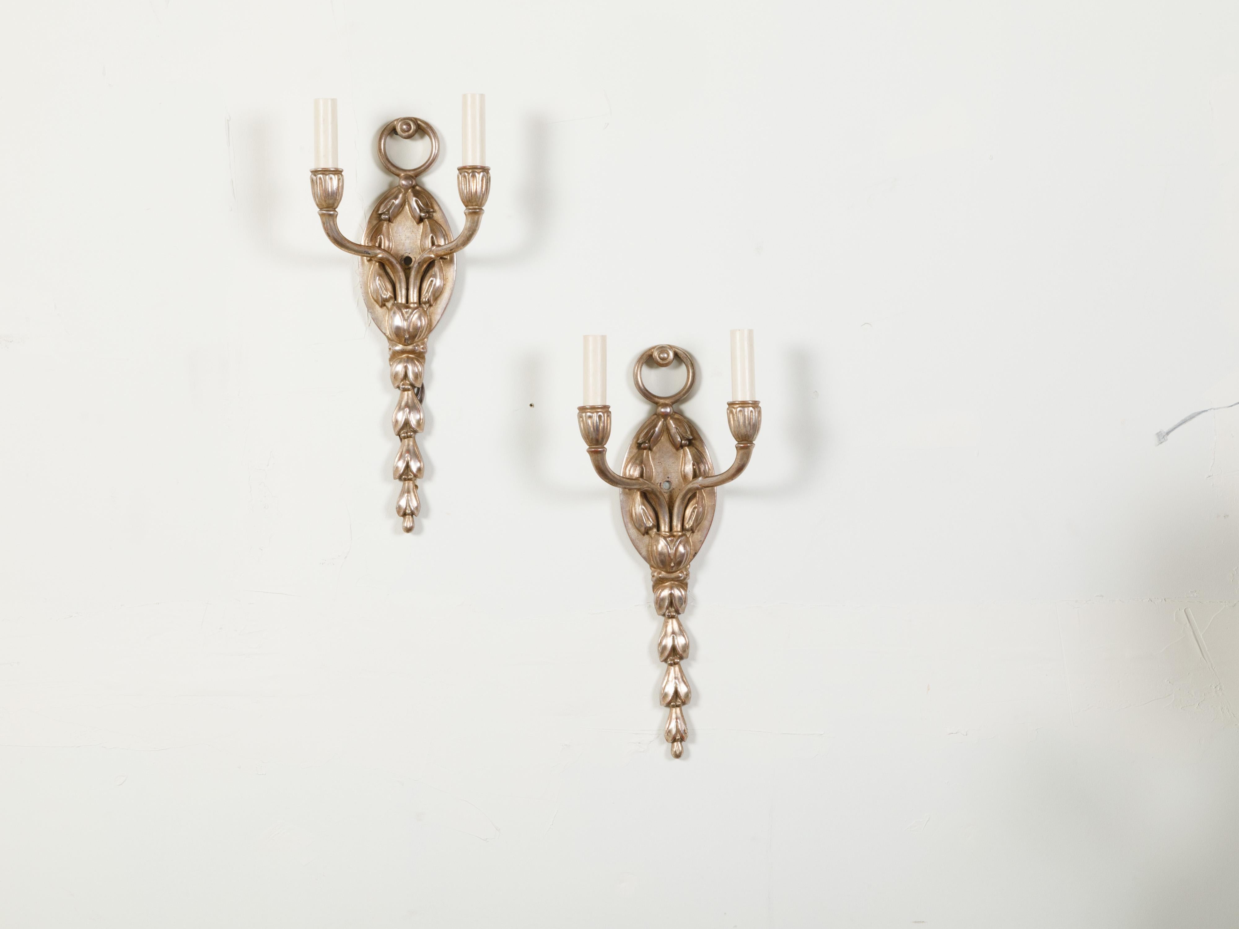 A pair of English vintage silver plated wall sconces from the mid 20th century, with two lights and foliage motifs. Created in England during the midcentury period, each of this pair of sconces, wired for the US, features an oval backplate adorned