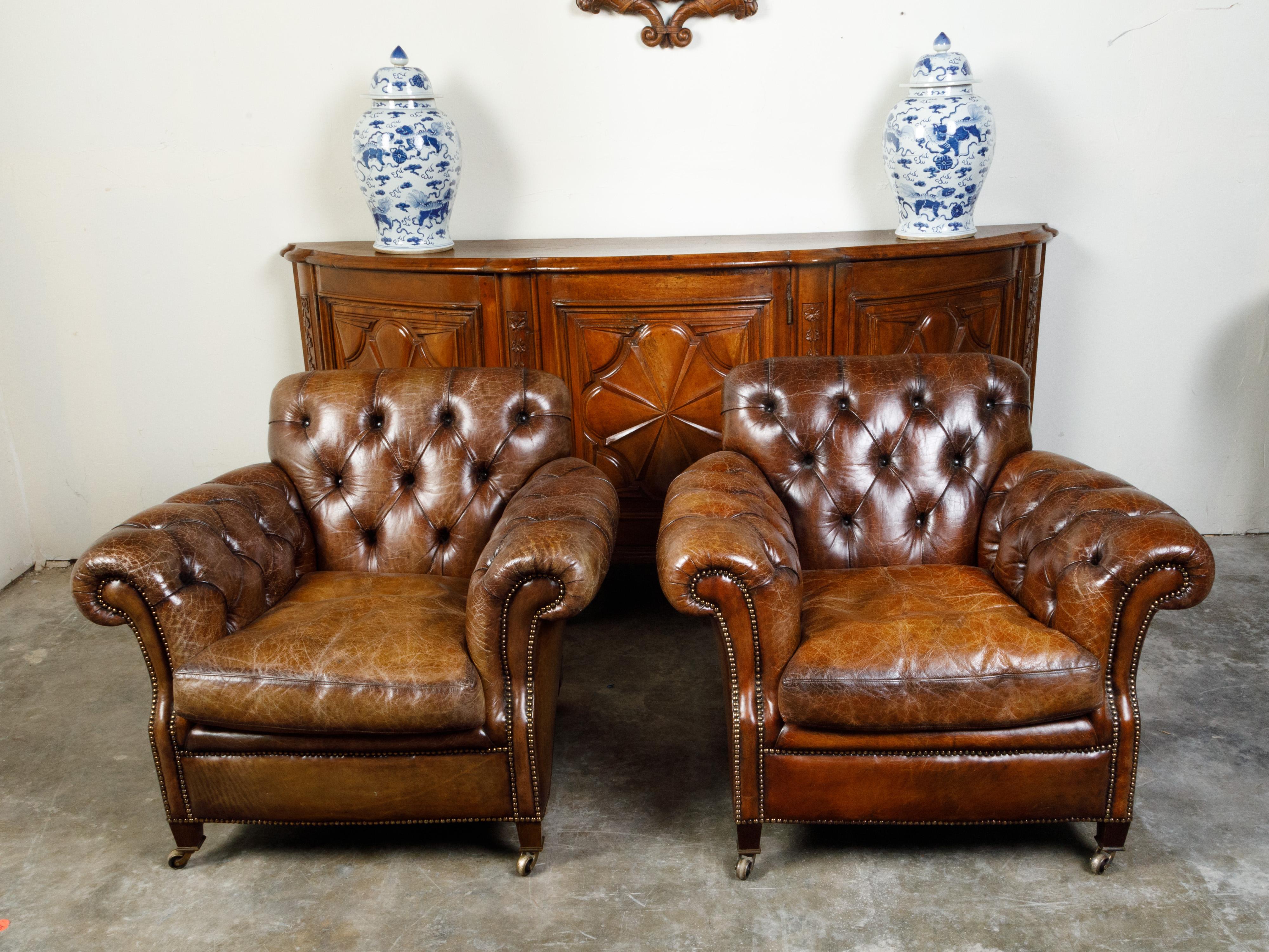 A pair of English vintage tufted leather club chairs from the mid 20th century, with out-scrolling arms and brass casters. Created in England during the Midcentury period, each of this pair of club chairs features a nicely weathered tufted leather
