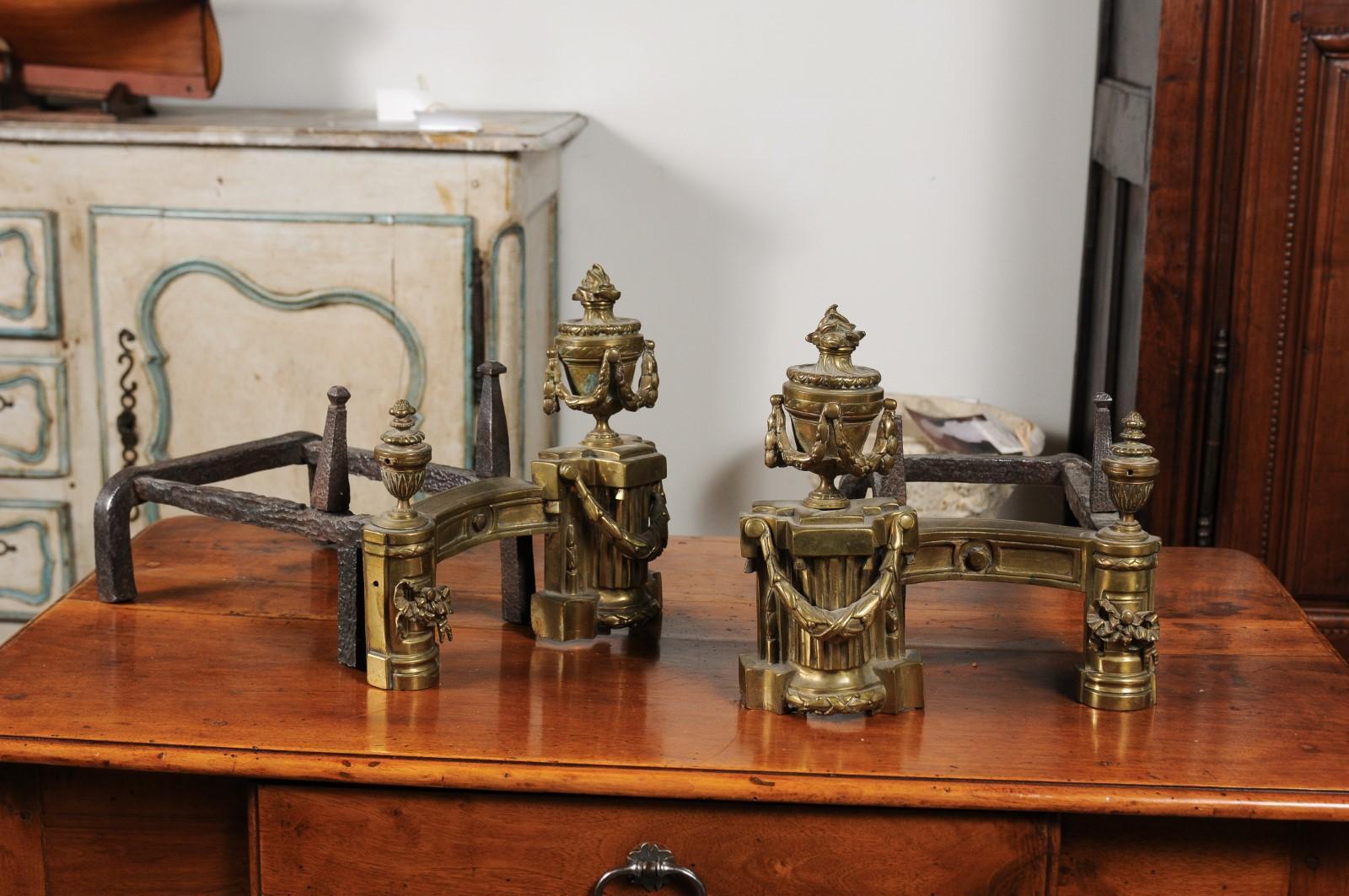 A pair of English neoclassical style brass andirons from the mid-19th century, with fire urns and swag motifs. Born in England during the reign of Queen Victoria, this pair of brass andirons features exquisite fire urns, standing proudly on