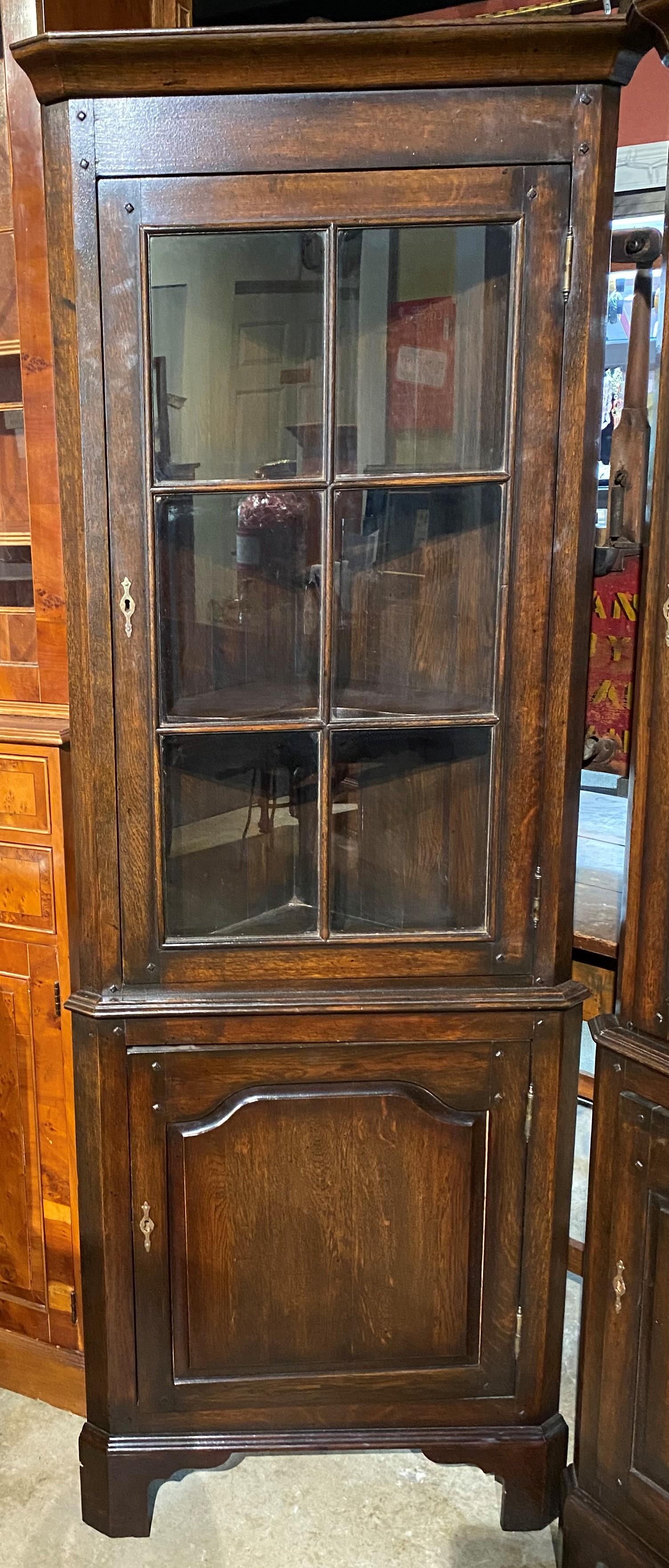 A diminutive pair of oak two door corner cupboards in their original dark finish, with molded cornice on each surmounting an upper door with six glass panels and brass escutcheon, opening to two interior shaped shelves, and a lower paneled door