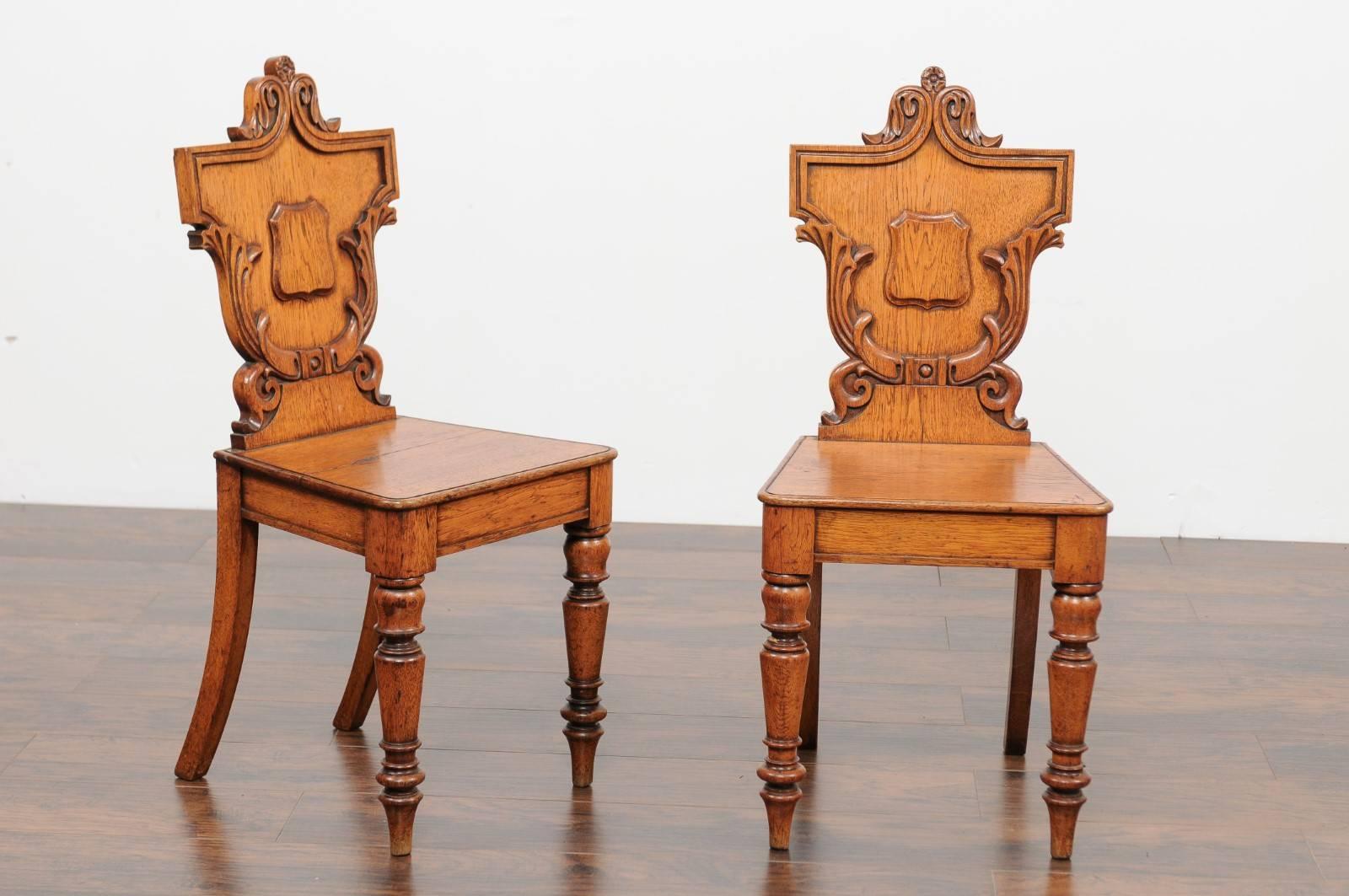 A pair of English oak hall chairs with carved shield-shaped backs from the late 19th century. Each of this pair of English hall side chairs features an exquisite back, skillfully carved with acanthus leaves surrounding a central cartouche while the