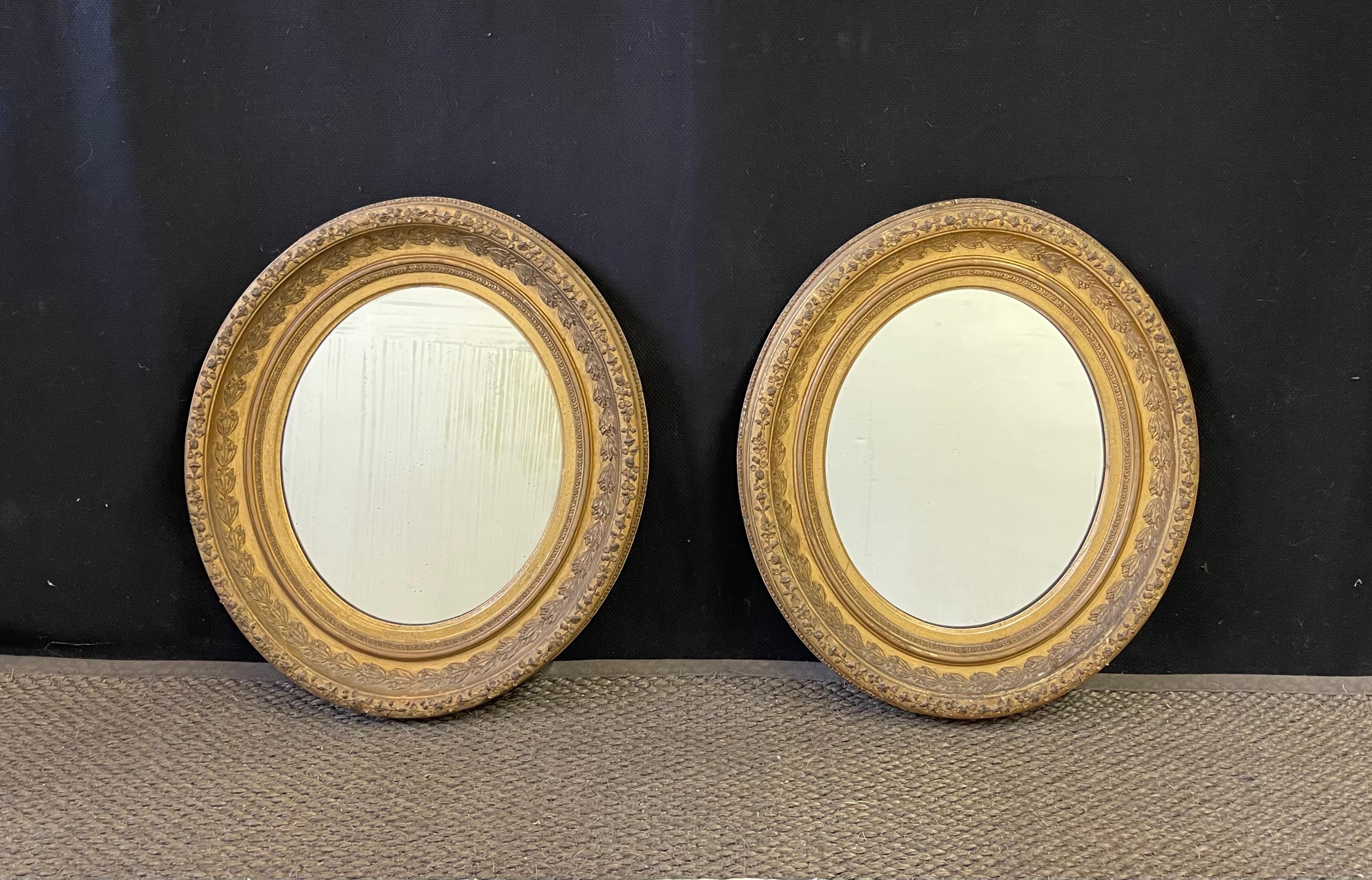 Lovely pair of 19th Century English gilt wall mirrors, oval in shape, and having bellflower, egg-and dart, and delicate floral trellis decorations on the frames. The antique mirrors are of the Victorian period and have the original glass which has