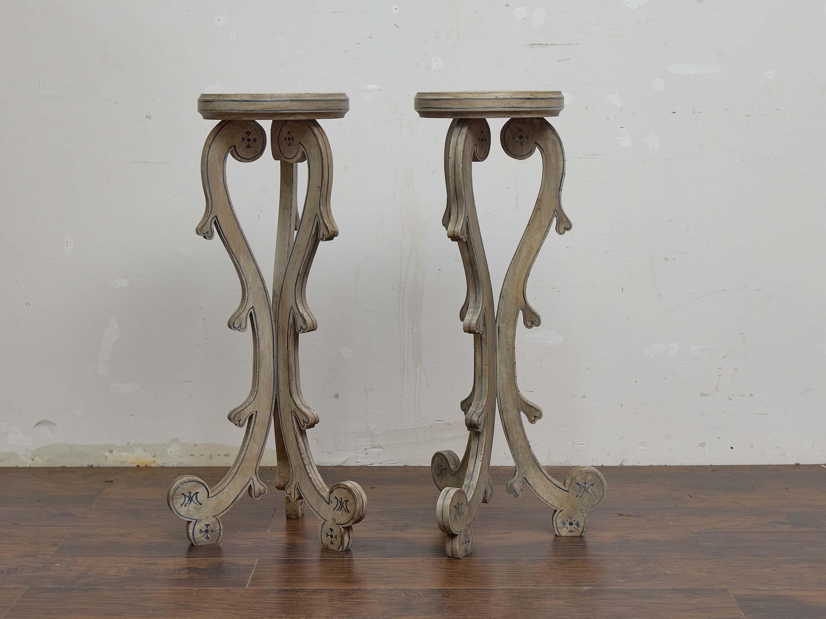 A pair of English painted pedestals from circa 1900, designed with elegant scroll-sawn supports and a light decorative finish. Each stand presents a circular top resting on three elongated legs with fluid curves, their ends rolled into scroll feet.