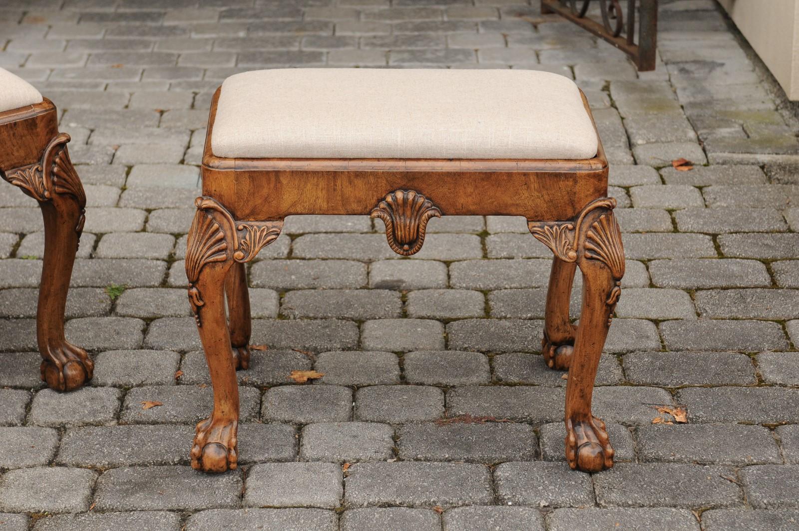 A pair of English Queen Anne style walnut stools from the early 20th century, with carved shells, ball and claw feet and new upholstery. Born during the first quarter of the 20th century, each of this pair of English Queen Anne style stools features