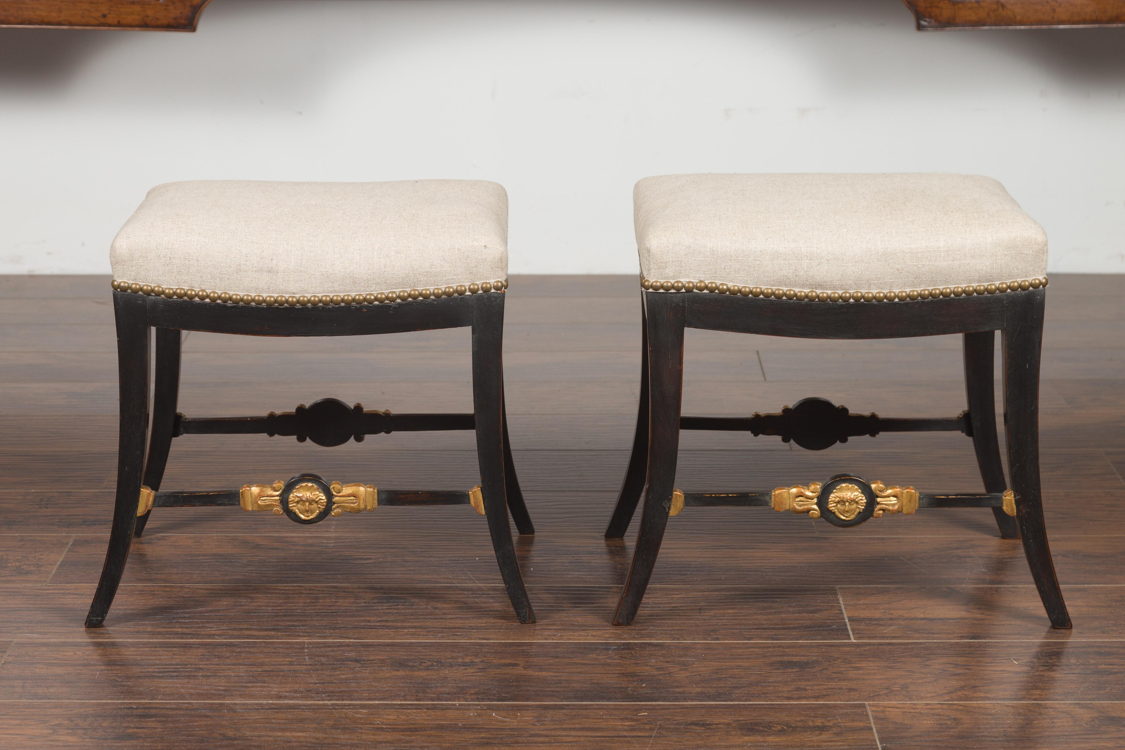 A pair of English Regency period ebonized and gilt stools from the early 19th century, with new upholstery. Created in England during the first quarter of the 19th century, each of this pair of Regency stools features a rectangular seat recently