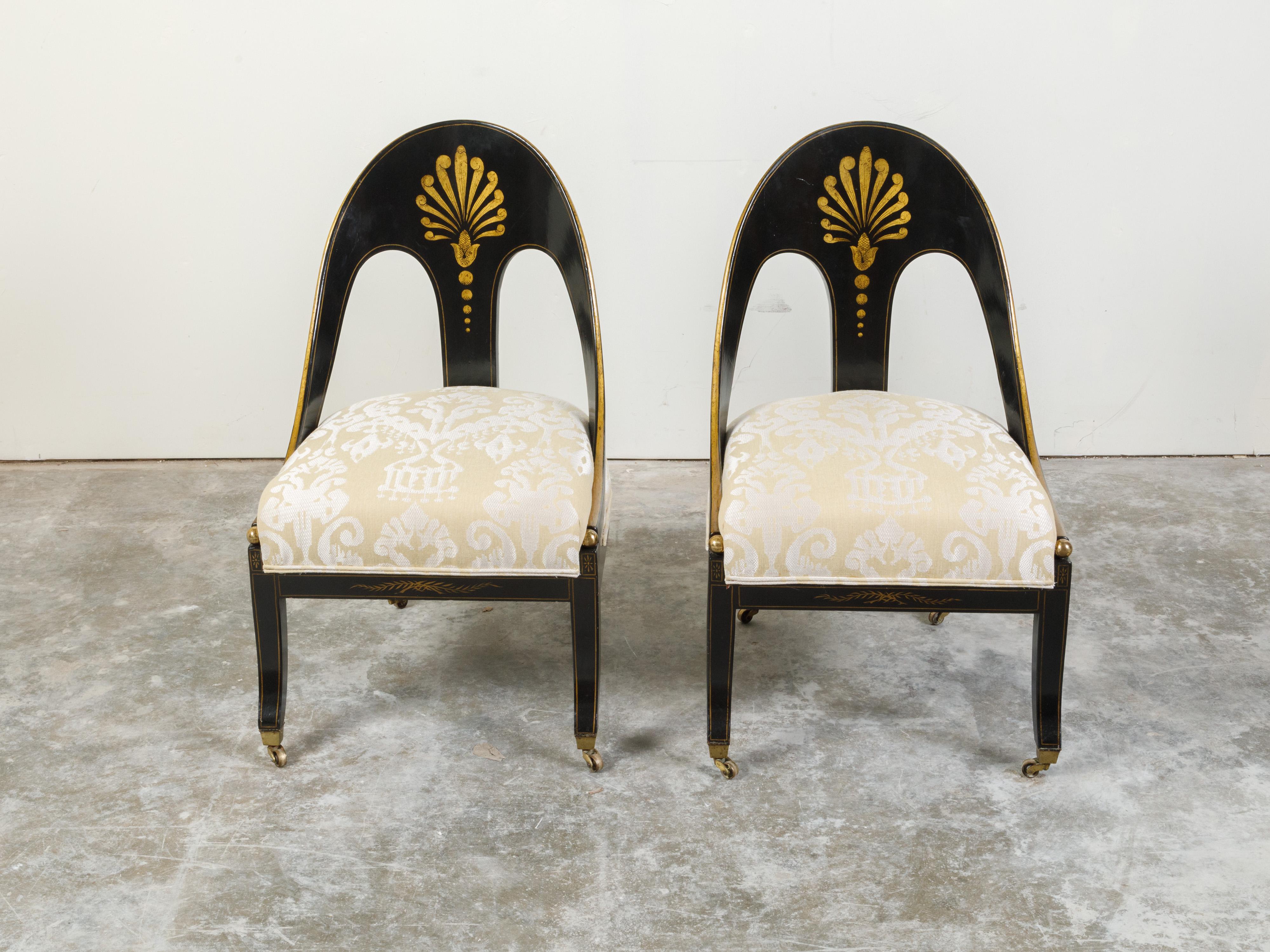 A pair of English Regency style black lacquer spoon back chairs from the 19th century, with gilt motifs and upholstery. Created in England during the 19th century, each of this pair of Regency style chairs features a spoon back beautifully adorned