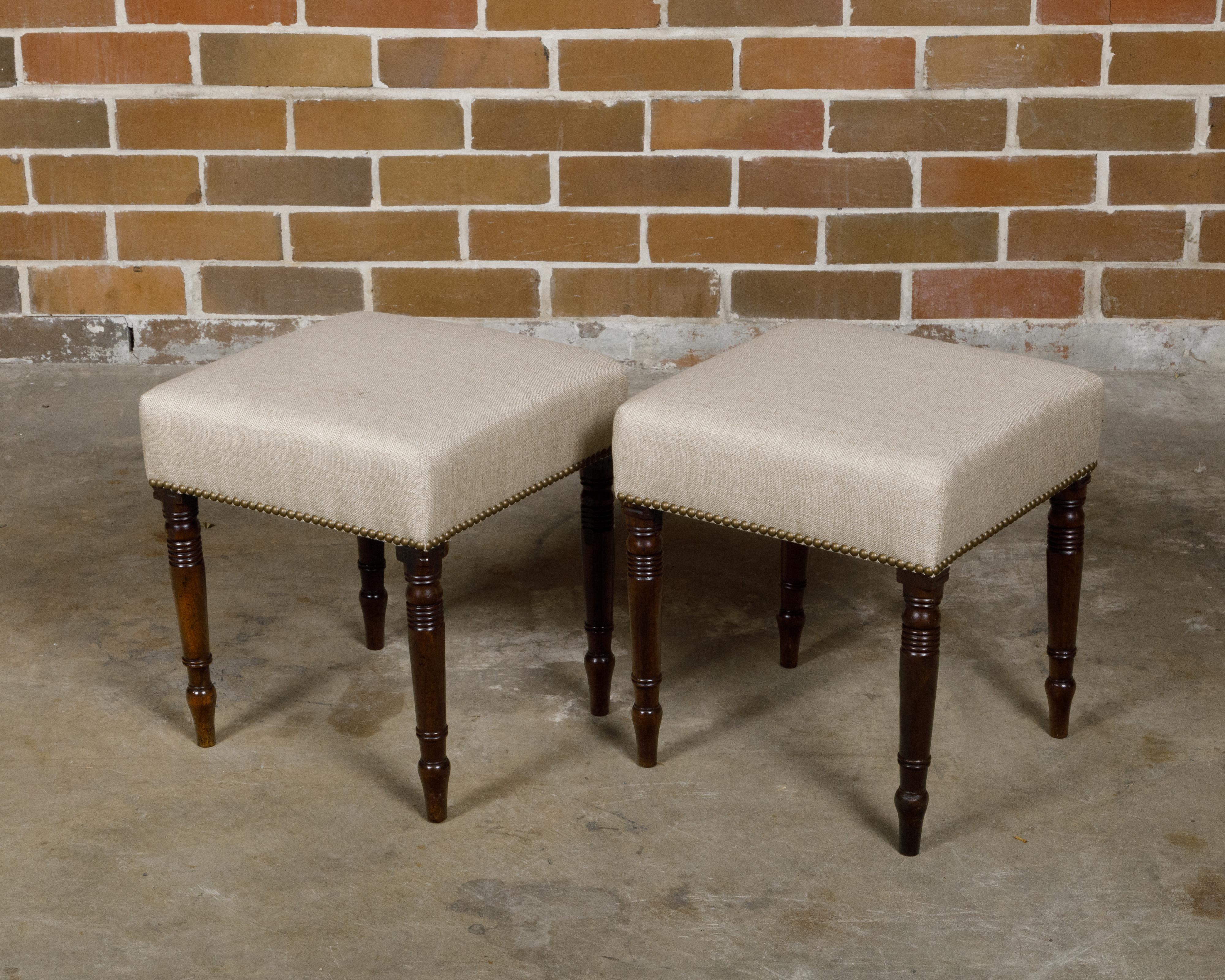 A pair of English Regency period mahogany stools from the 19th century with turned spindle shaped legs and linen upholstery. This pair of English Regency period mahogany stools, dating back to the 19th century, marries the elegance of classic design