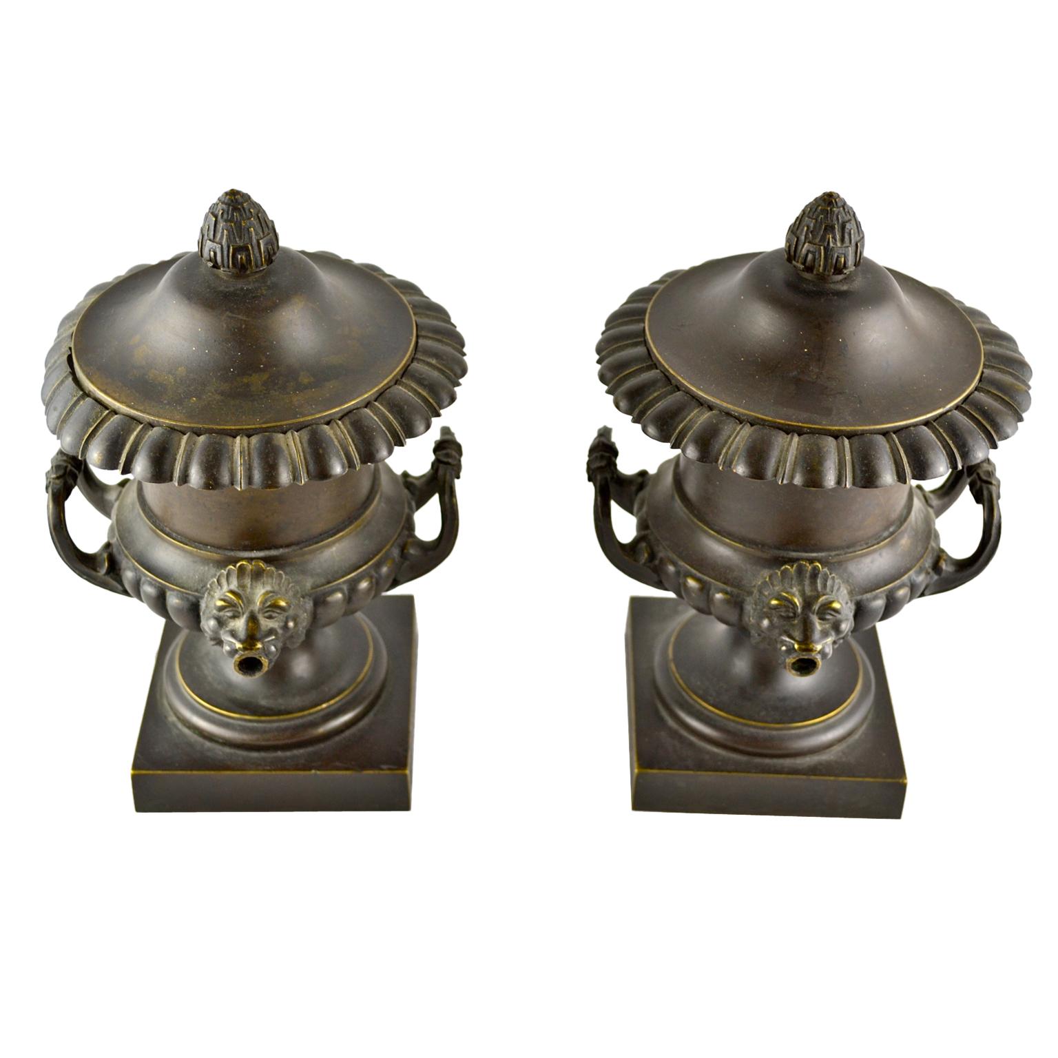 A pair of English Regency bronze perfume burners in the classical campana urn style, the urns with acorn finial topped lids and lion’s head spouts around a middle egg and dart rim from where perfume was diffused into the room. One of the burners has