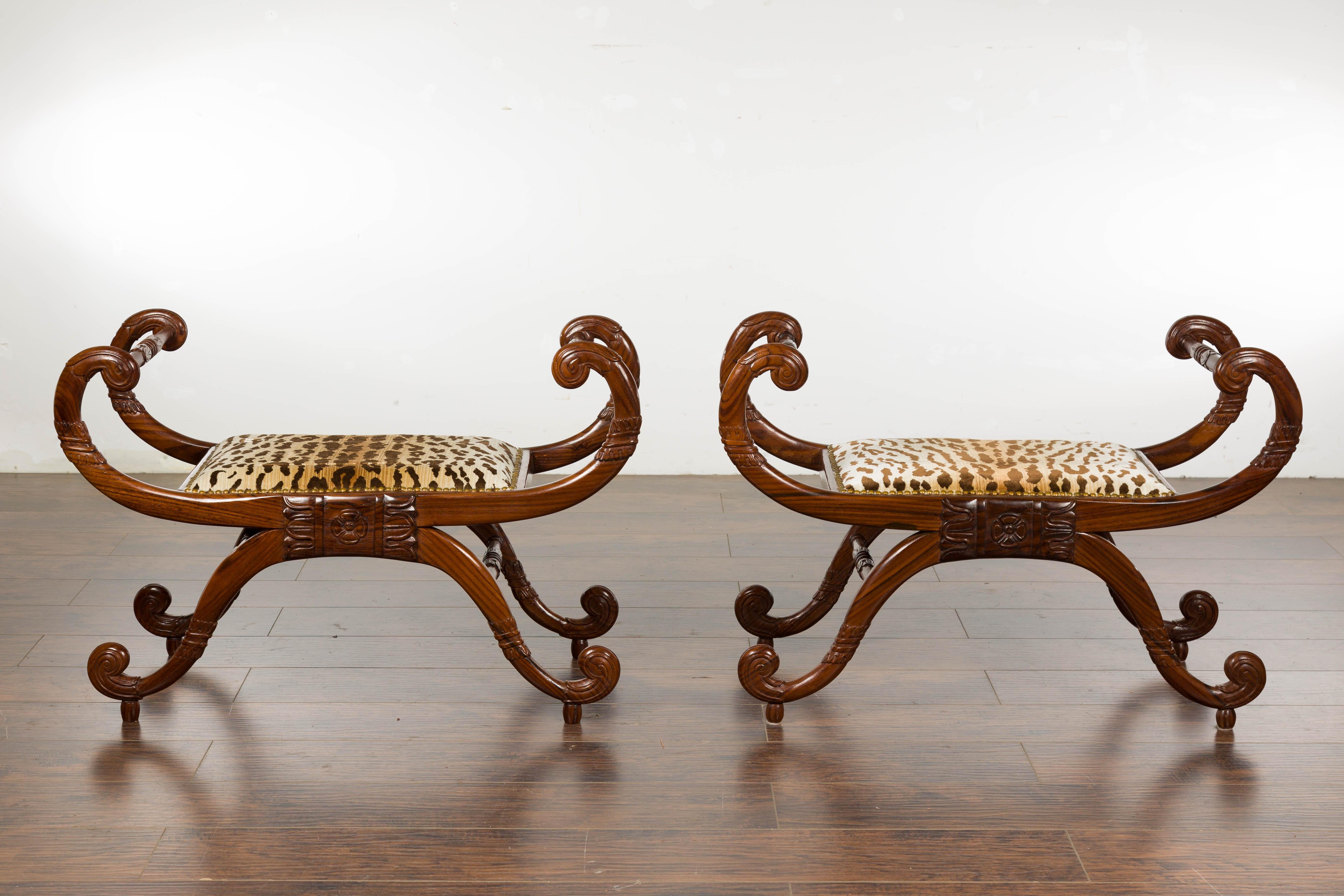 A pair of English Regency mahogany stools from the 19th century with large scrolling arms and legs, carved foliage and tiger style upholstered seats. Imbue your home with the distinguished charm of the 19th-century English Regency period with this