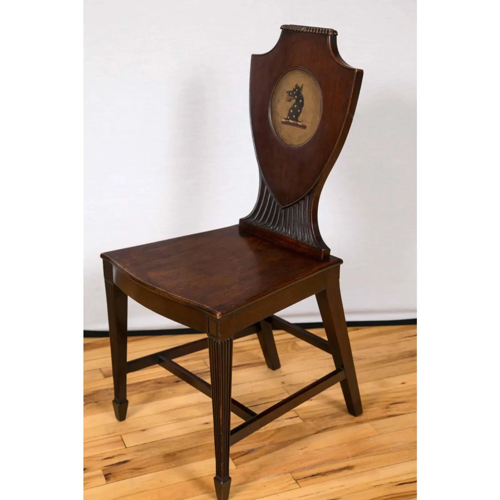 A pair of late 18th century mahogany hall chairs with shield form backs centered by oval reserves painted with an heraldic devices. Bow front seats are raised on tapered and reeded legs, terminating in spade feet.