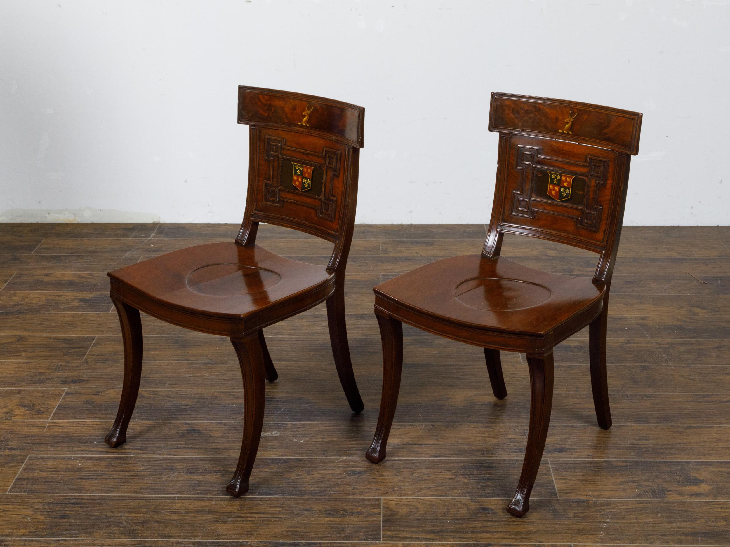 A pair of English Regency mahogany hall chairs from the 19th century with coat of arms, dished seats and saber legs. These English Regency mahogany hall chairs from the 19th century are a testament to the elegance and refinement of the period.