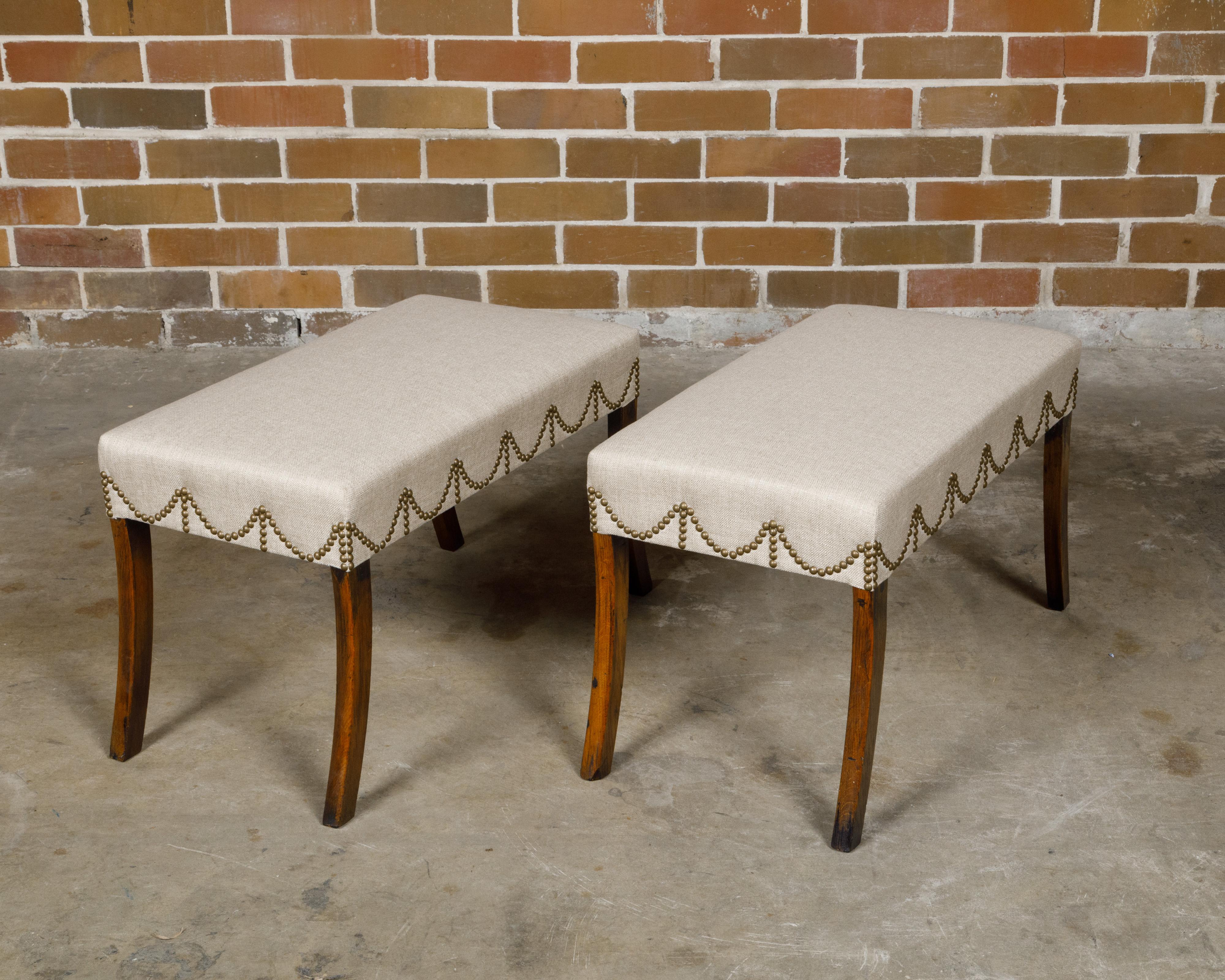 A pair of English Regency period mahogany rectangular stools from the 19th century with saber legs and custom upholstery. This exquisite pair of English Regency period mahogany stools from the 19th century is a testament to the era's elegance and