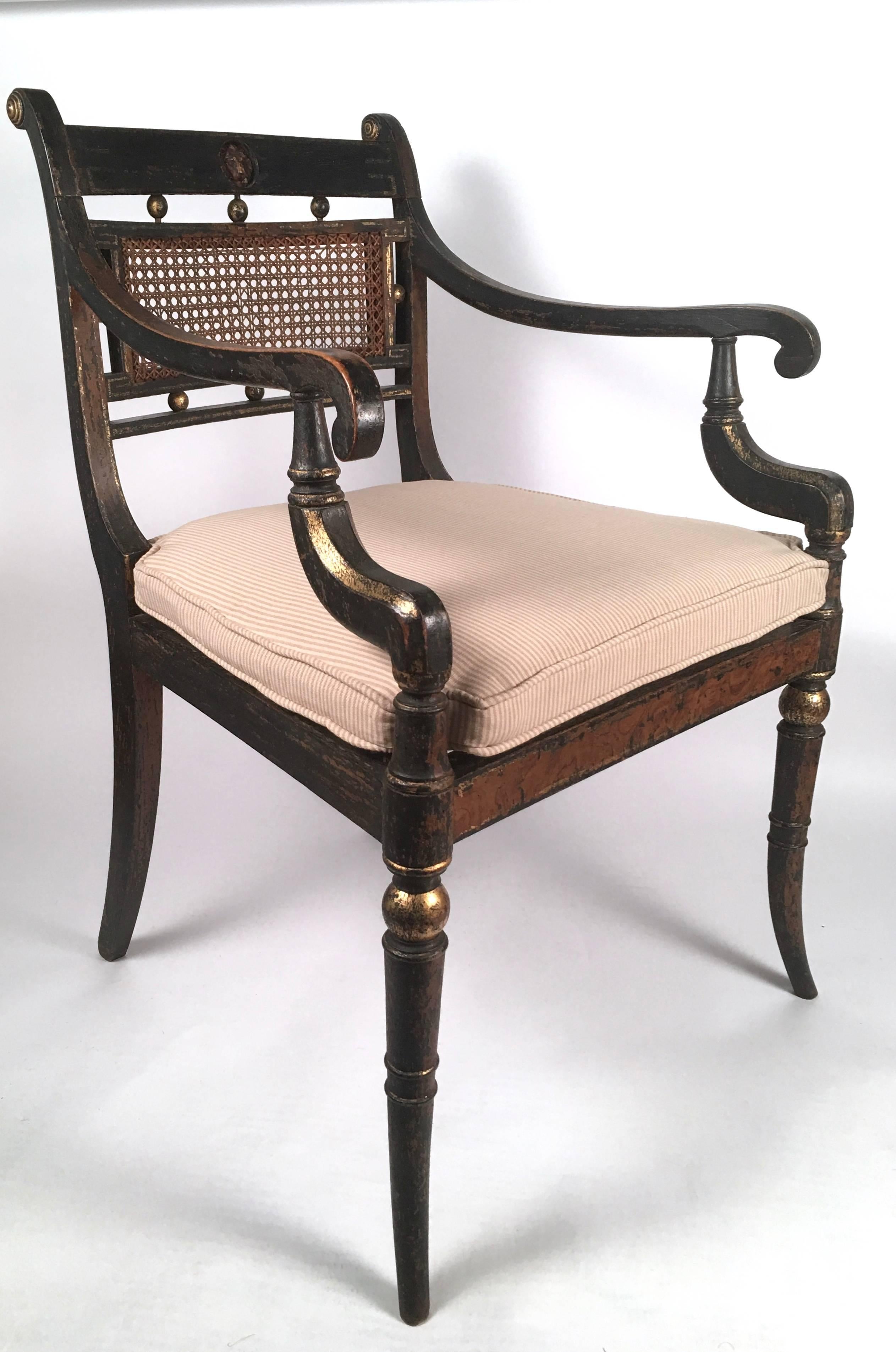 A pair of elegant, well proportioned English Regency period ebonized and parcel gilt open arm chairs, with carved lions masks on the backs above gilded spheres and a caned back, flanked by elegantly scrolled downswept arms over a conforming caned