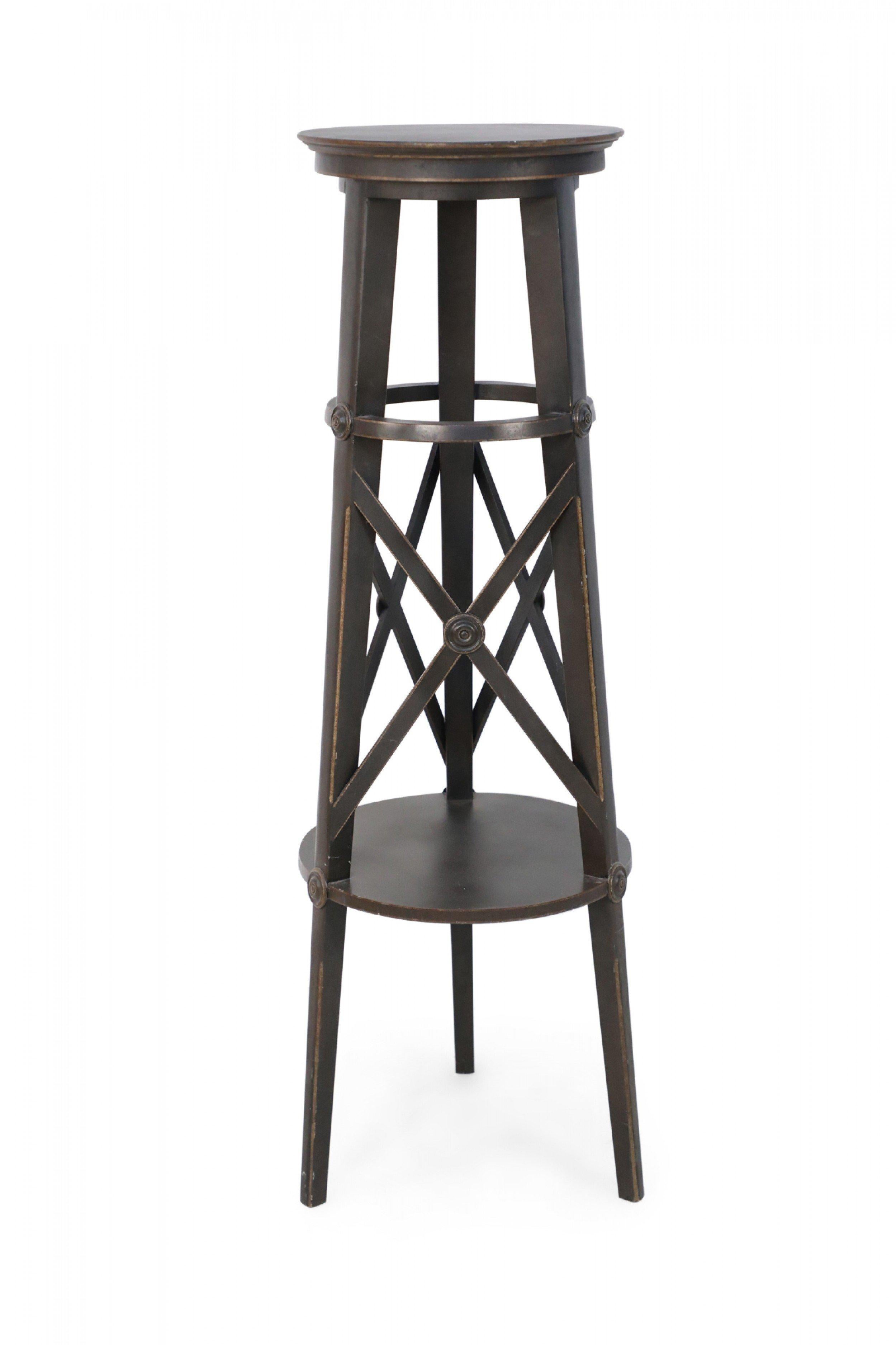 Pair of tall circular brown painted wooden pedestals with top and bottom shelves connected by three legs with x-shaped supports, featuring carved circular medallions to emphasize where the lines intersect. (priced as pair).