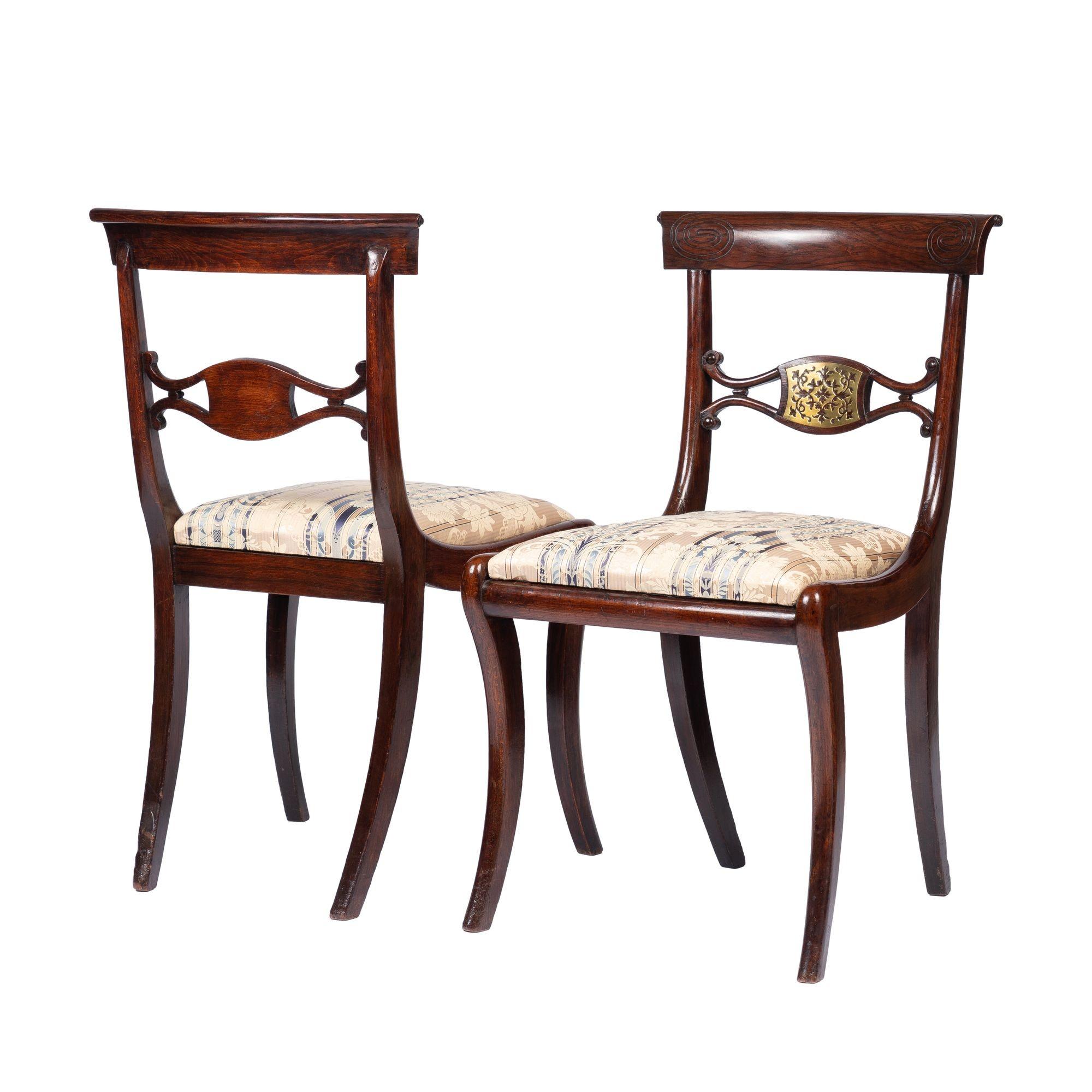 Pair of English Regency Period faux rosewood painted beechwood Klismos side chairs inlaid with pierced brass embellishments on the center of the back rail and the corners of the crest rail. The chairs are fitted with upholstered slip seats on fine