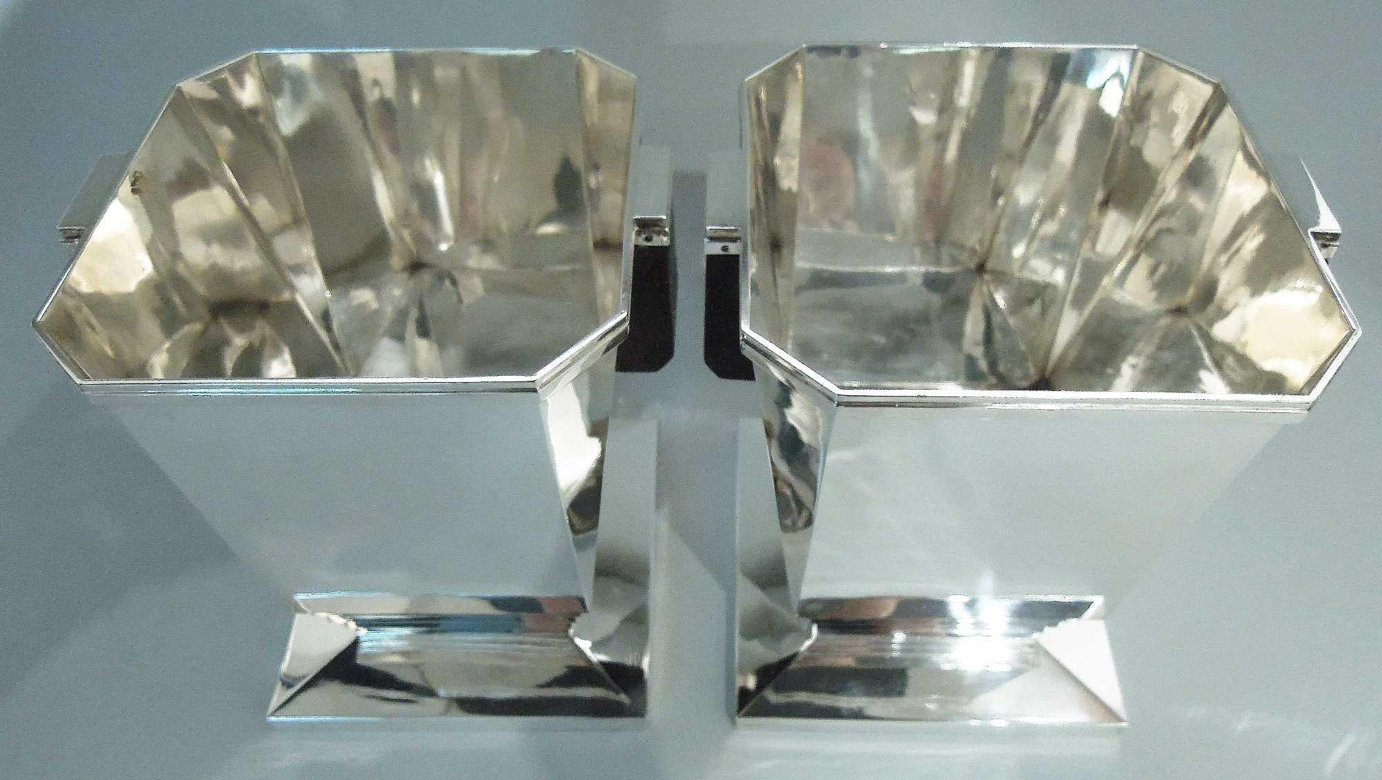 Pair of English Retro Deco sterling silver wine coolers, 2014. Each: Chamfered and tapering bowl; stained-wood side handles with stepped sterling silver mounts. Tapering and stepped support mounted to chamfered square base. All planes and angles for