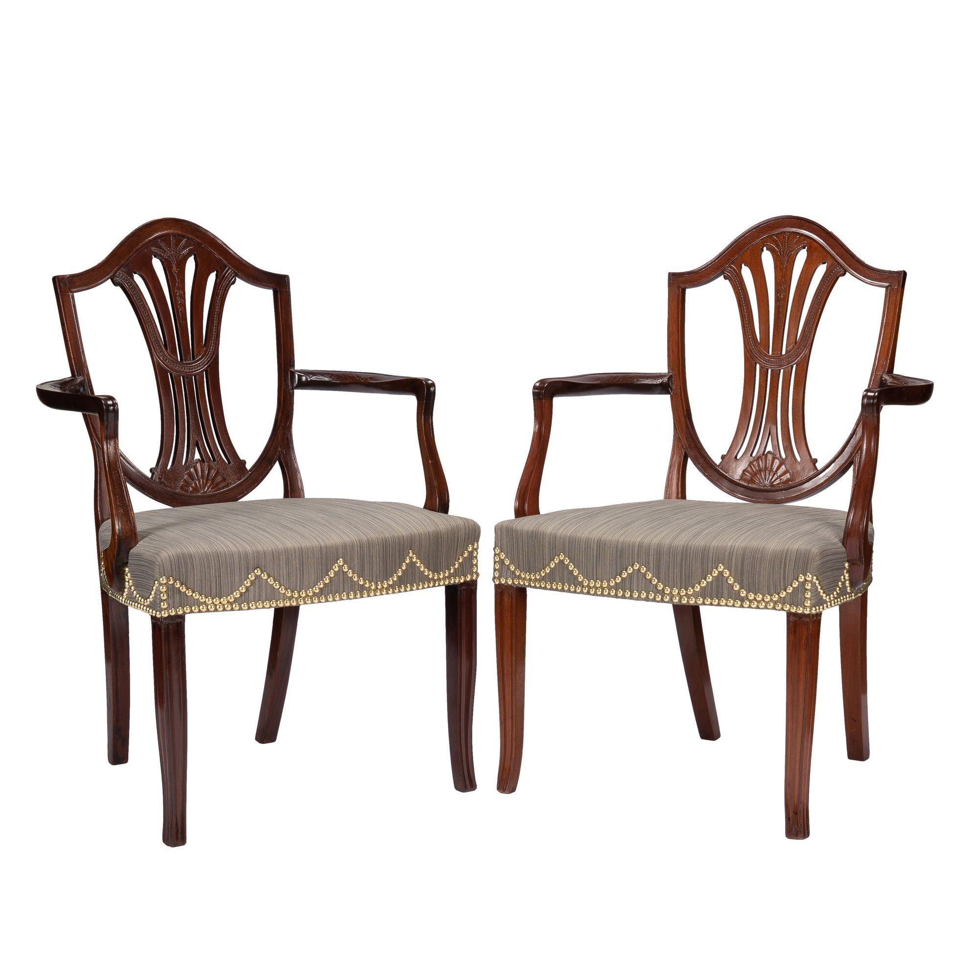 Pair of generously proportioned English Sheraton mahogany shield back arm chairs with over rail upholstered seats. The chair backs are detailed with a finely carved rice sheaf above a bell flower in the center of the vertical back splat. The elbow
