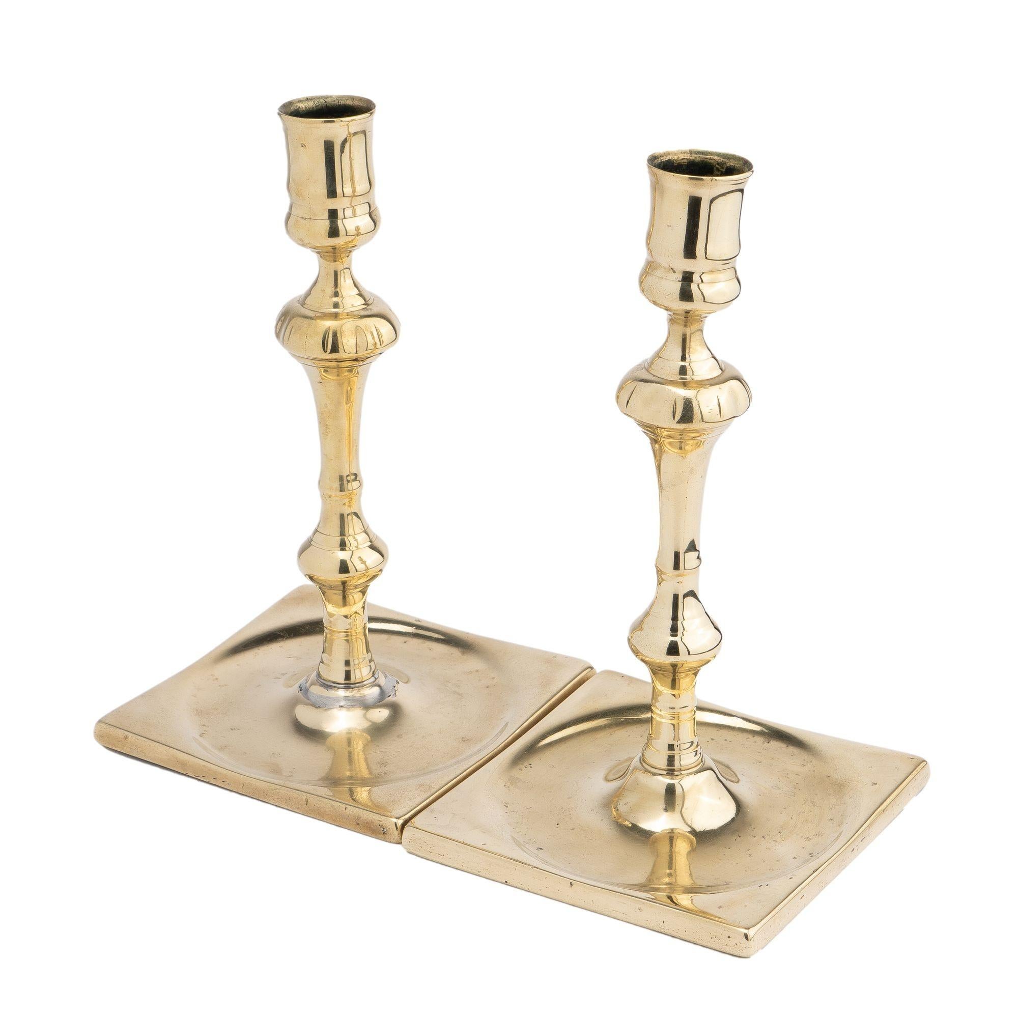 Pair of silver form core cast brass baluster candlesticks with square bases. The knobbed candle shaft is peened to the center of a suppressed drip pan within the square base. The candle shaft supports a classic urn form candle cup, without a