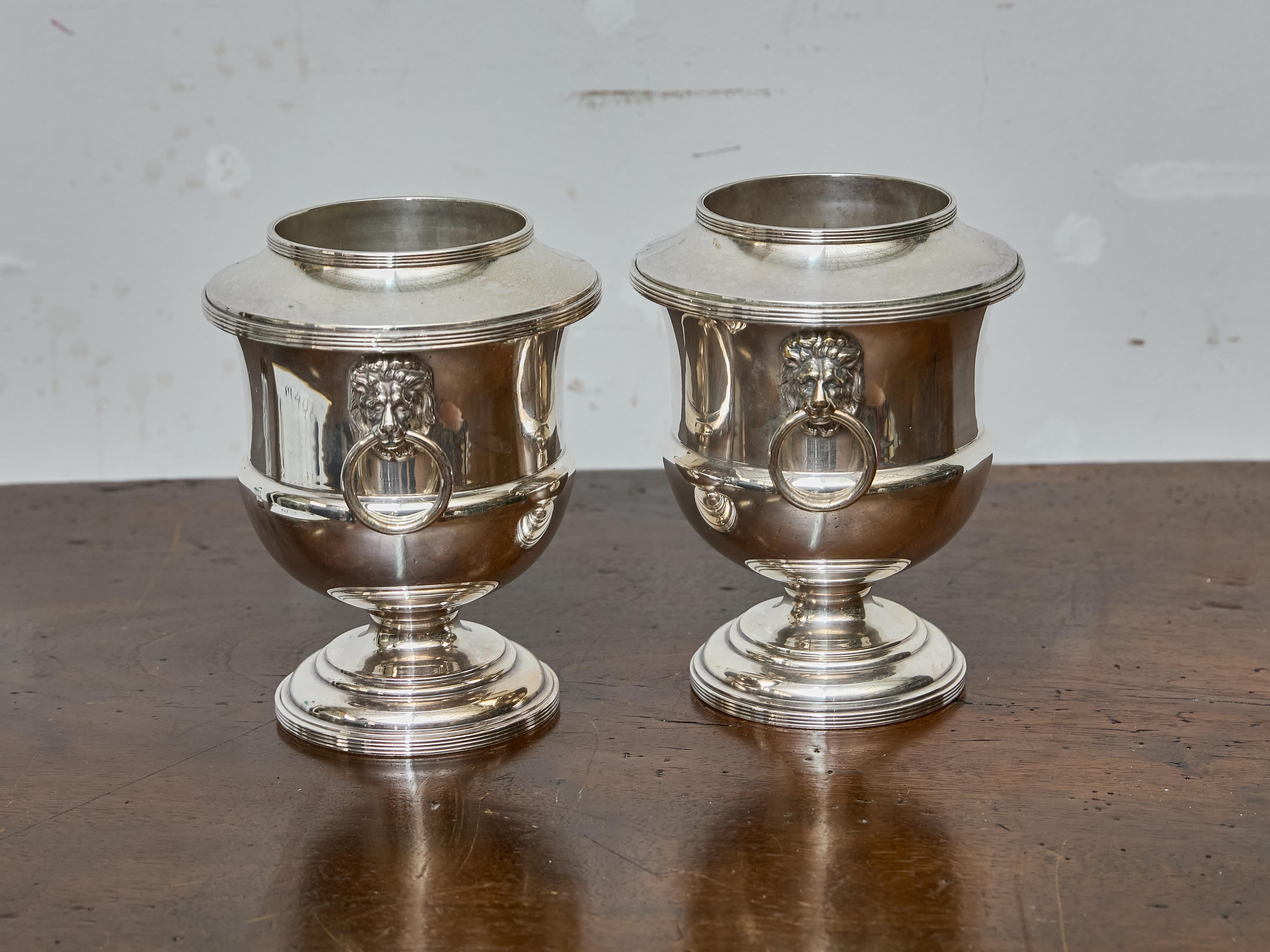 An elegant pair of English silverplate wine coolers from circa 1900, each featuring a classic urn form raised on a circular pedestal base. The coolers are distinguished by their refined neoclassical design and beautifully cast lion mask handles,