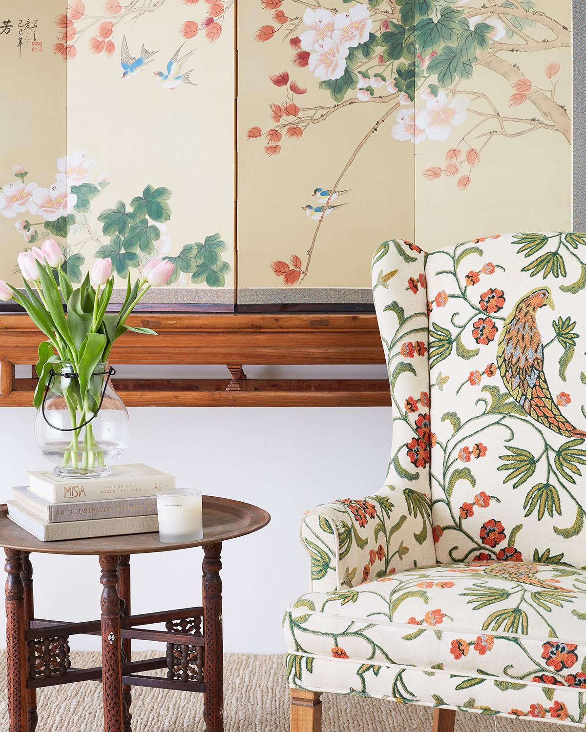 Stunning pair of English style wing chairs or wingback chairs featuring an upholstered crewel work frame. The crewel work embroidery depicts a large peacock style bird among brightly colored flowers and foliate vine designs. Each chair back is