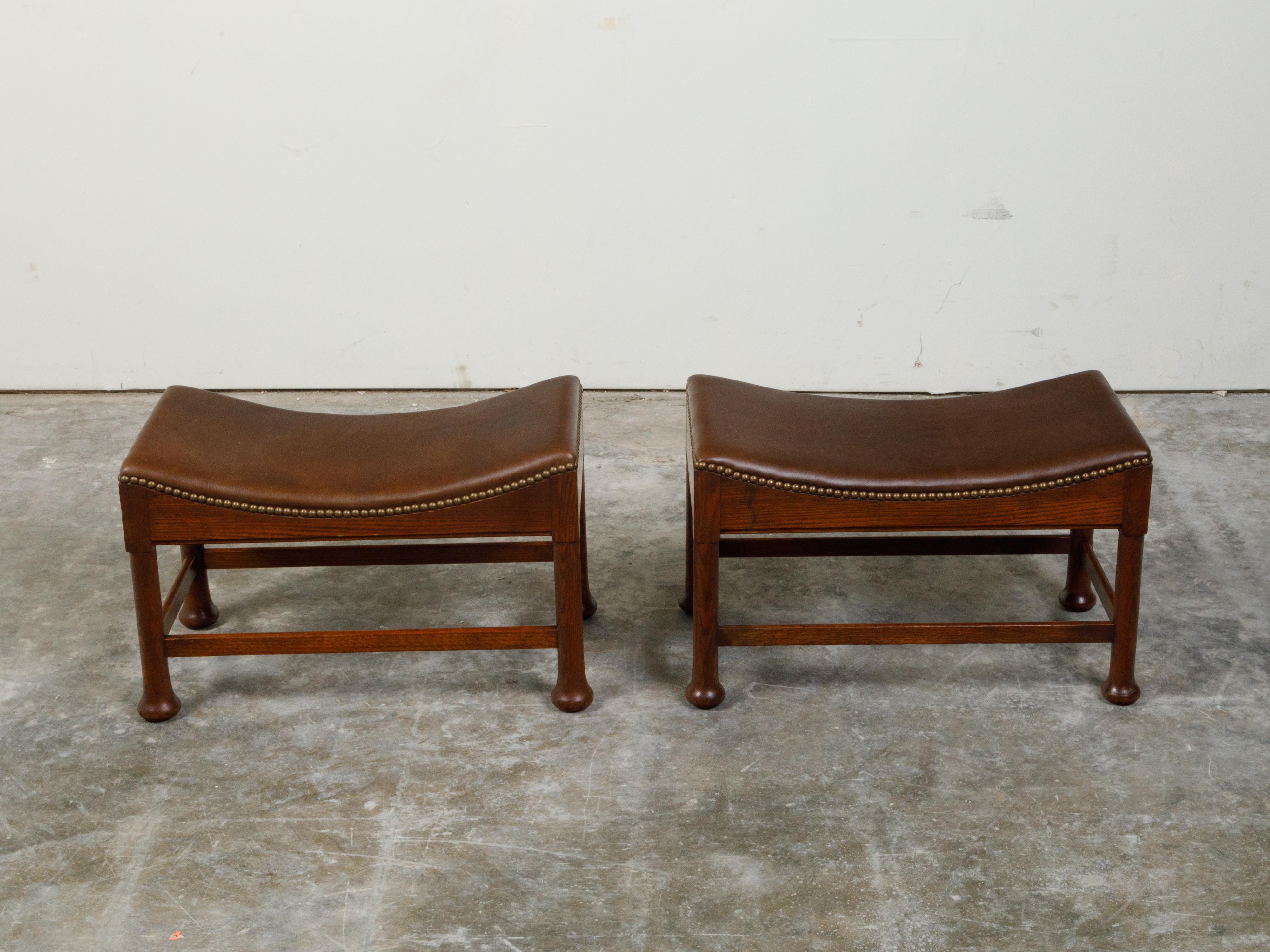 A pair of English oak stools from the early 20th century, with curving seats and pad style feet. Created in England at the Turn of the Century, each of this pair of oak stools features an in-curving upholstered seat with brass nailhead, sitting