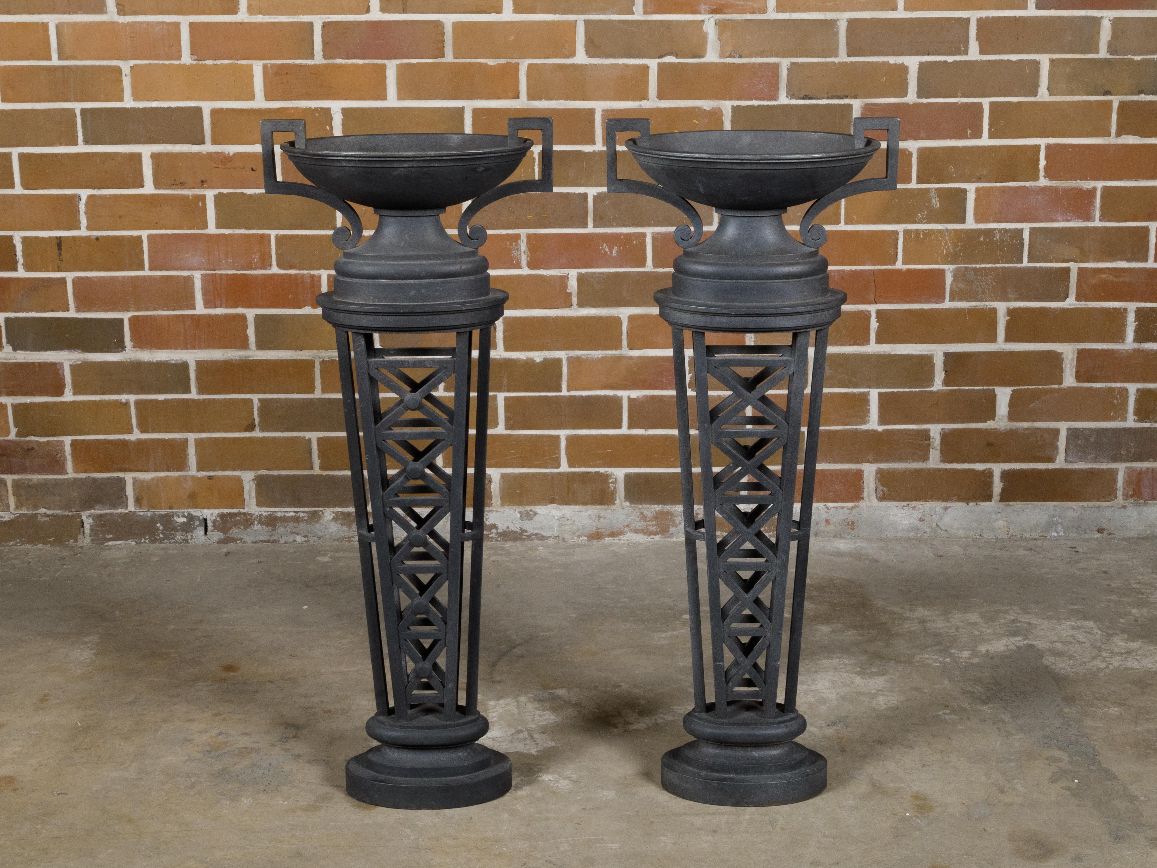 A pair of English Victorian period cast iron planters from the 19th century with Neoclassical urns sitting atop tapering pilasters. This exquisite pair of English Victorian period cast iron planters, dating back to the 19th century, beautifully