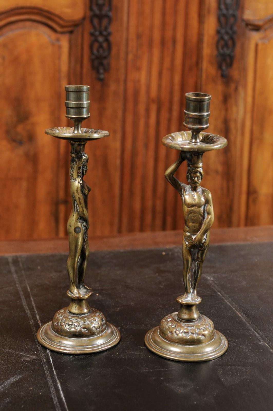 A pair of English Victorian period Adam and Eve brass candlesticks from the 19th century, on circular bases. Created in England during the reign of Queen Victoria, this pair of brass candlesticks features a man and woman, likely Adam and Eve