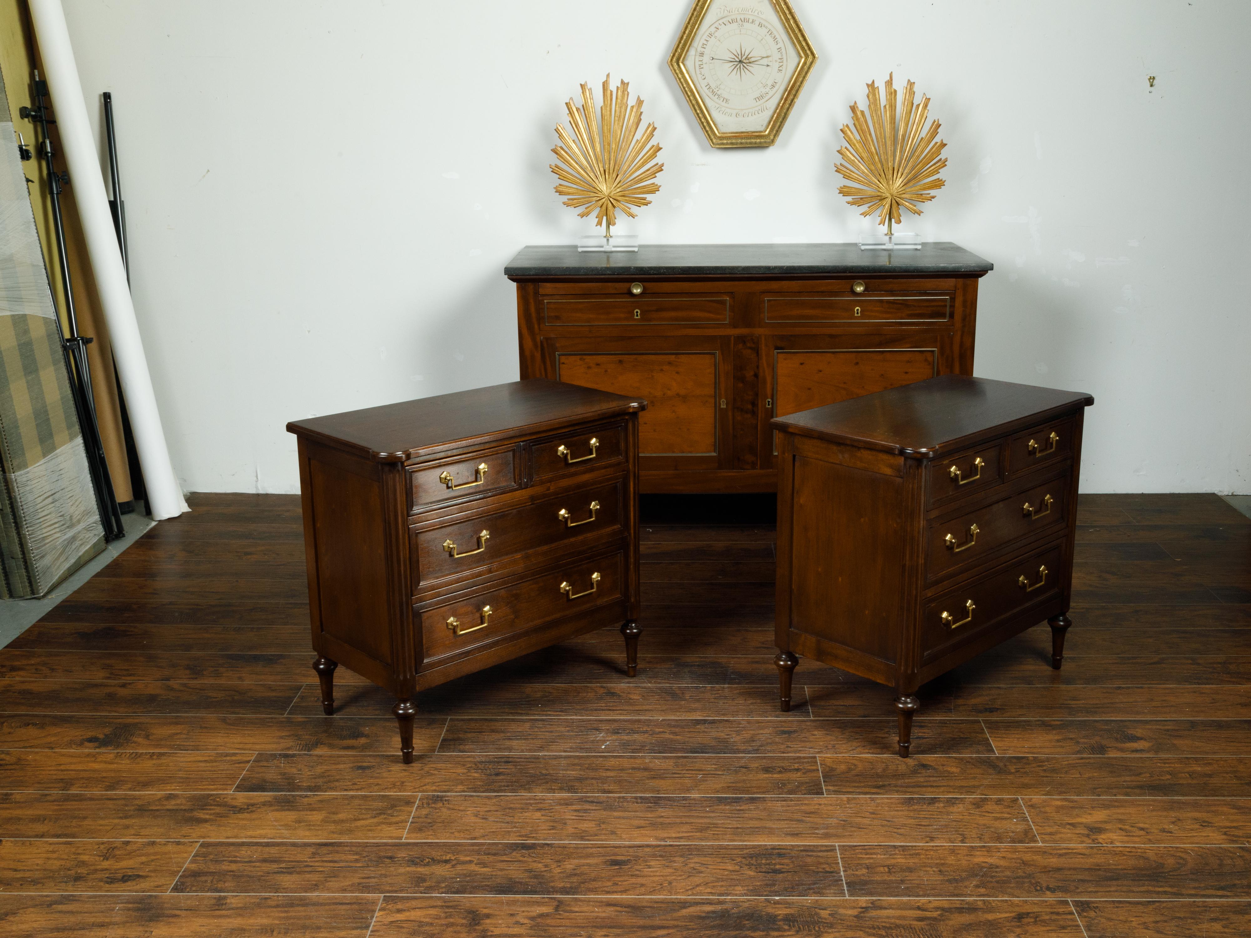 A pair of English mahogany commodes from the mid 20th century, with four drawers, brass hardware and fluted side posts. Created in England during the mid 20th century, each of this pair of commodes features a rectangular top with rounded protruding