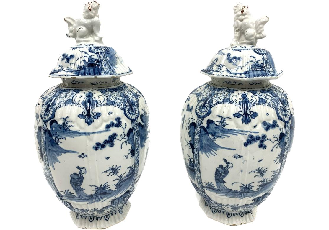 Pair of blue and white footed ginger jars with foo dog finials. The ginger jars with figural chinoiserie decorations within cartouches and flow from one panel to the next bordered by geometric bands. The jars with raised fluted motifs around the top