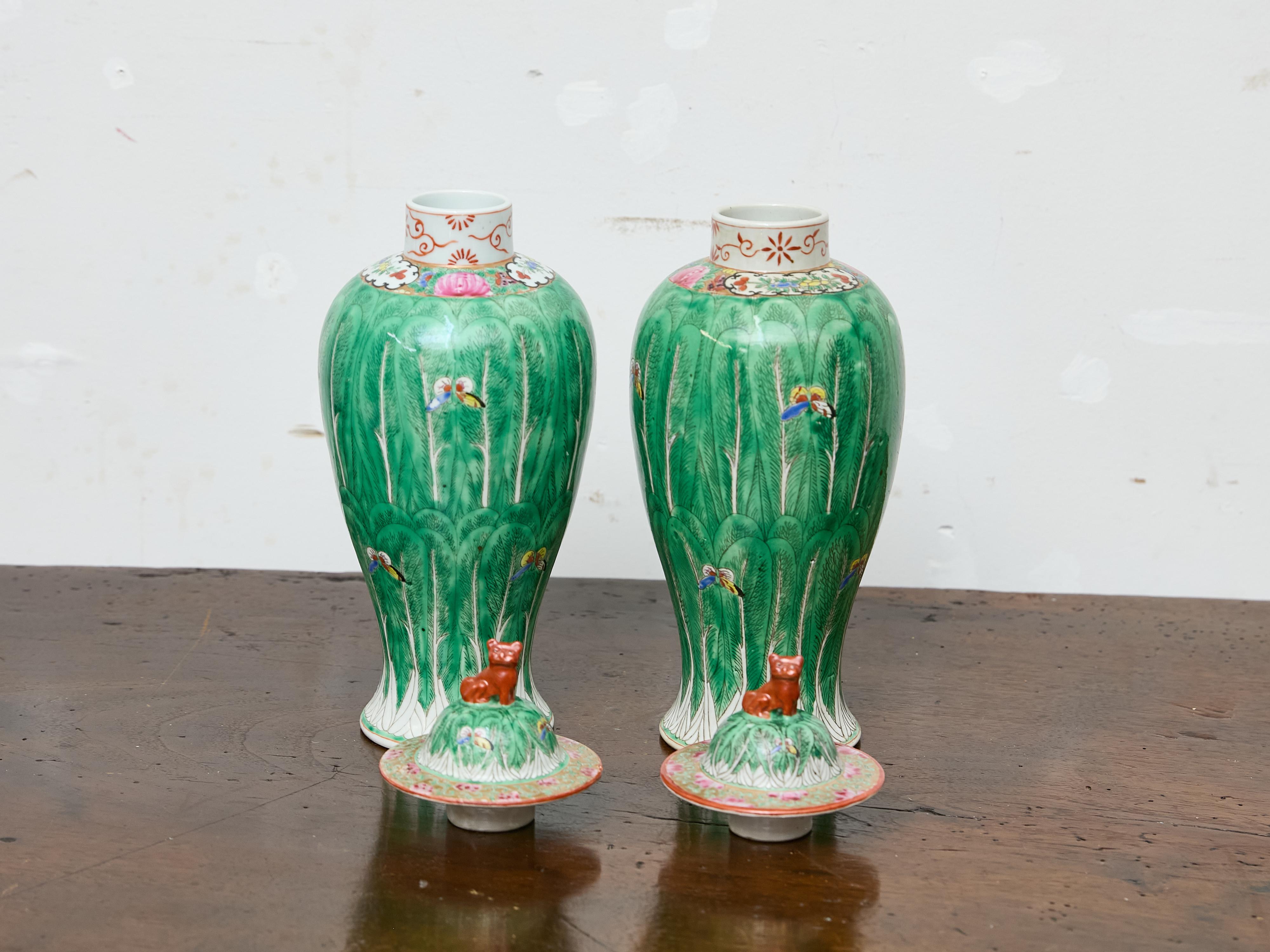 This decorative pair of Chinese famille verte porcelain vases dates to circa 1920 and exemplifies the richly colored aesthetic of early 20th-century export ceramics. Each baluster-form vase is topped with a domed lid featuring a red-brown foo lion