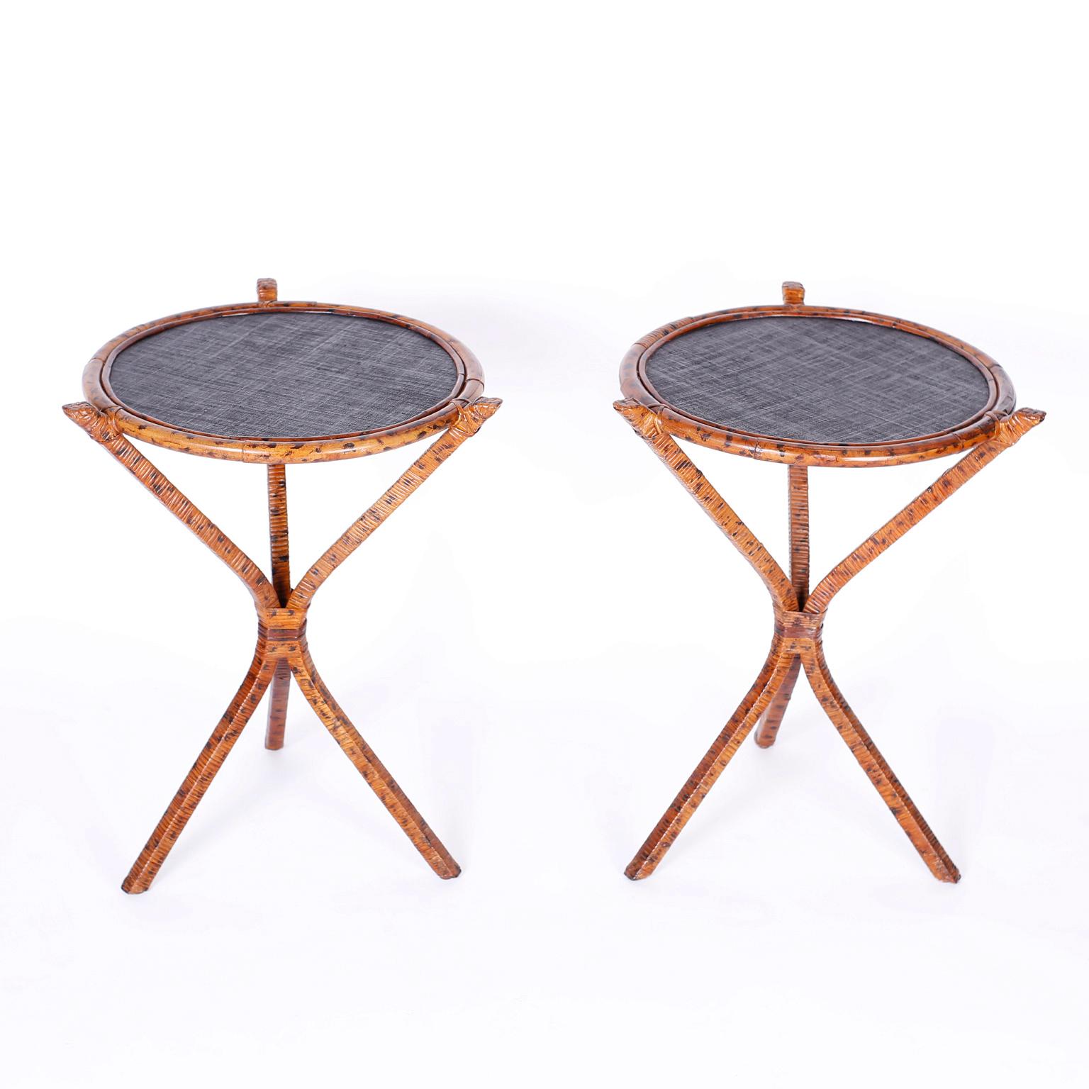 Pair of British colonial style tables with round black grasscloth tops framed with burnt bamboo on three legged metal bases wrapped in reed and decorated in faux burnt bamboo.