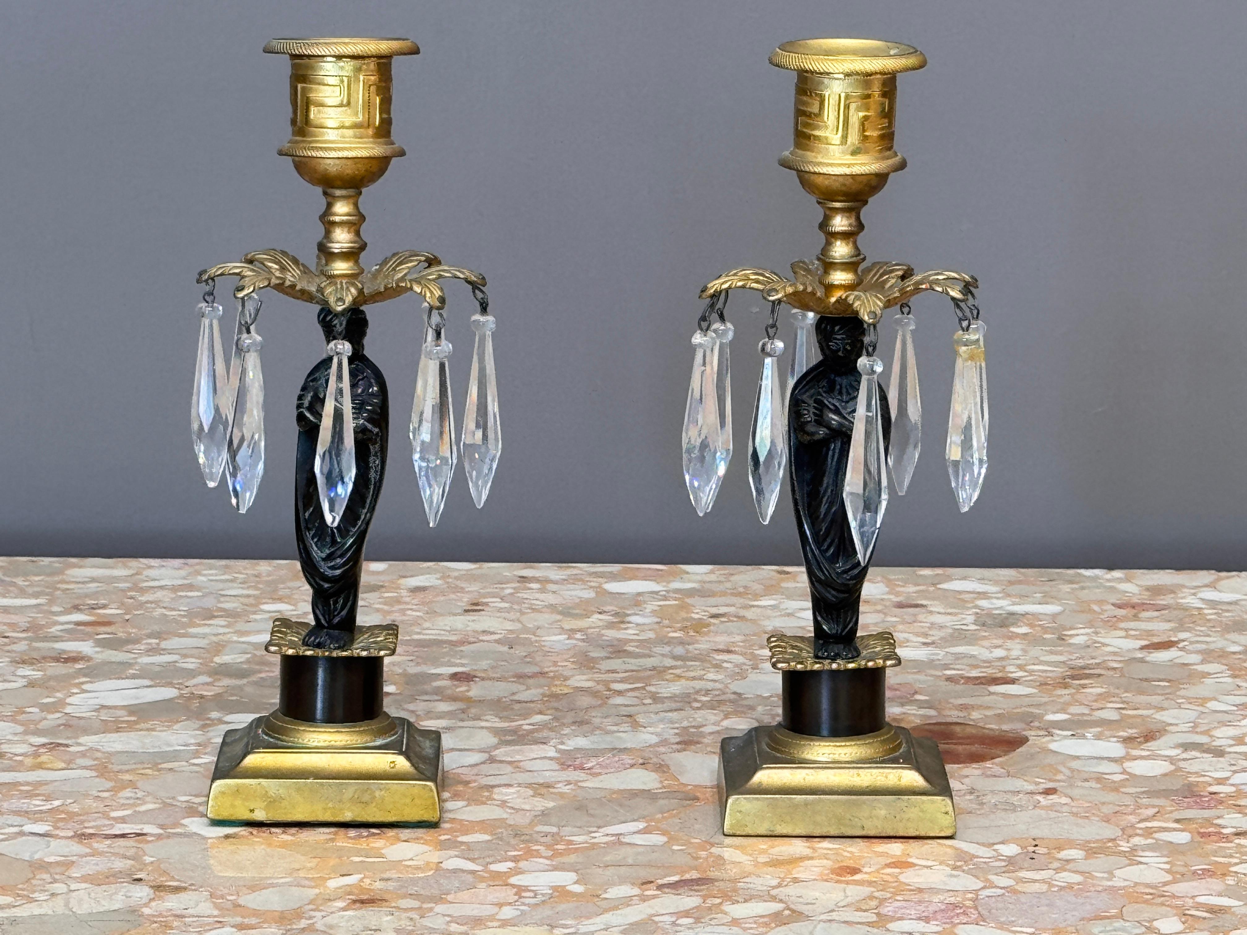 A finely detailed pair of figural girandoles in ormolu and patinated bronze, each modeled as a classical draped figure supporting a single candle cup. The gilt nozzles feature Greek key decoration and are set over leafy bobeches with suspended