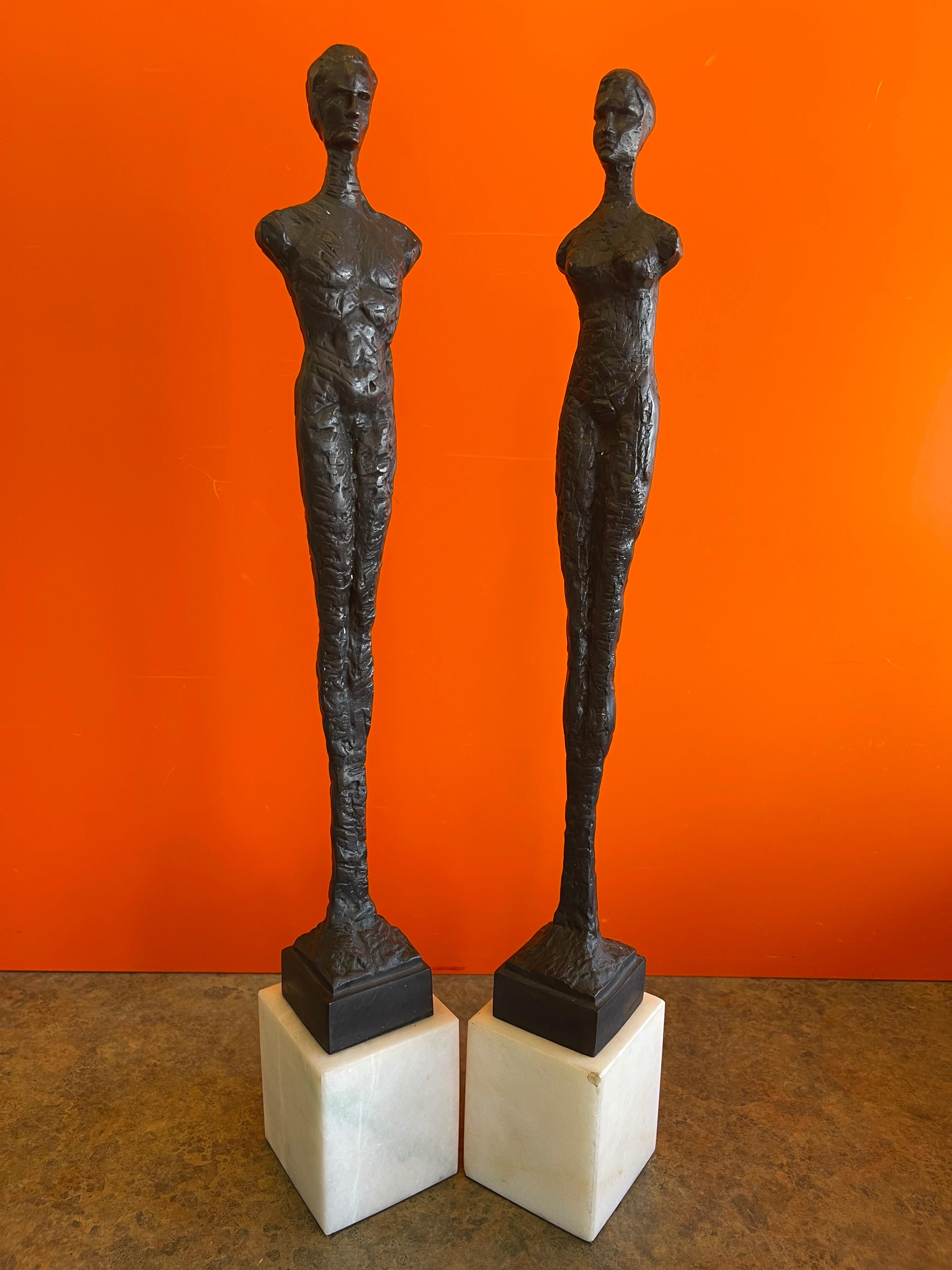 Pair of figurative bronze man & woman sculptures in the style of Giacometti, circa 1970s. The bronzes sit on white marble bases that measure 3