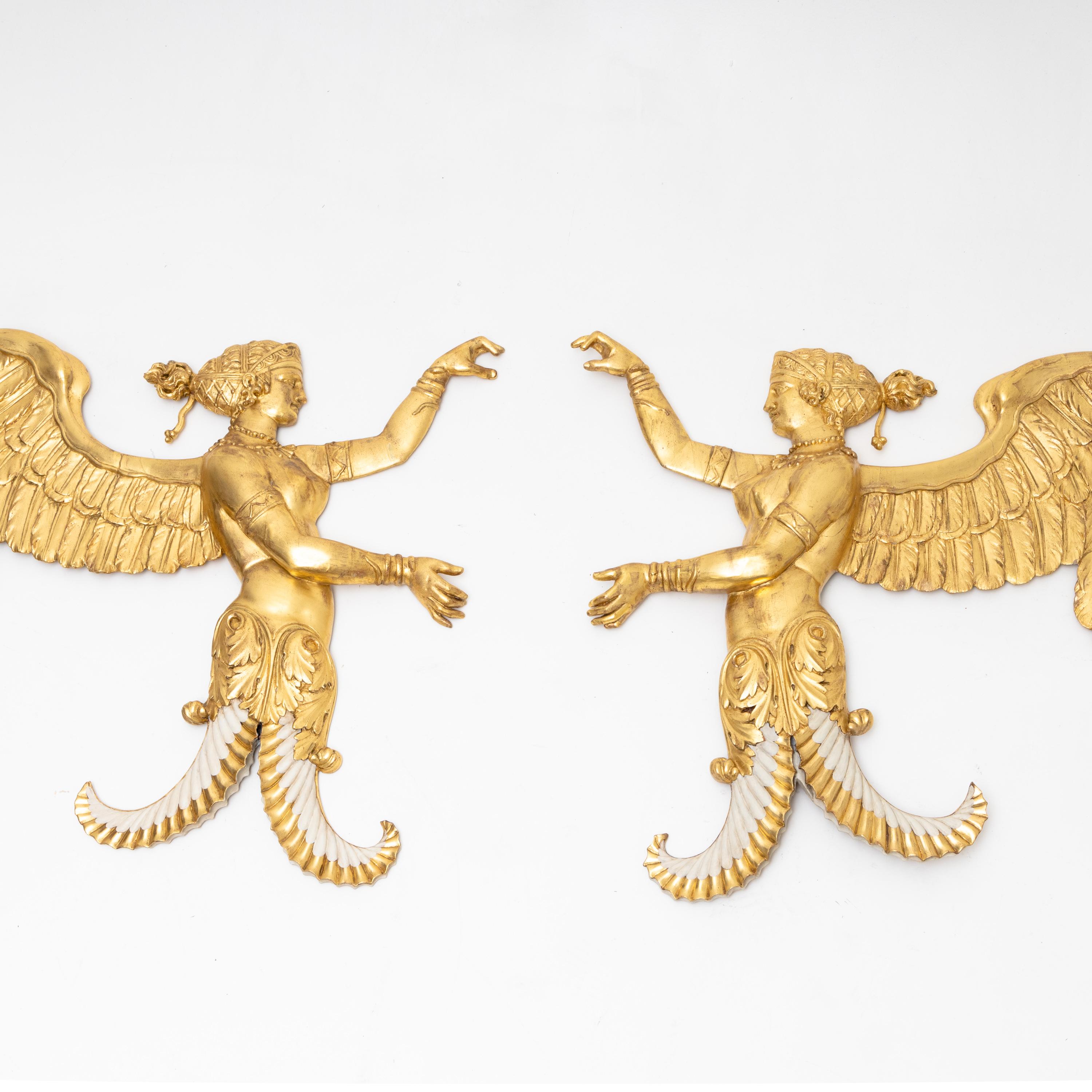 Pair of gold patinated wall reliefs in the form of winged female half-figures with detailed decoration and arms raised in a framing gesture.