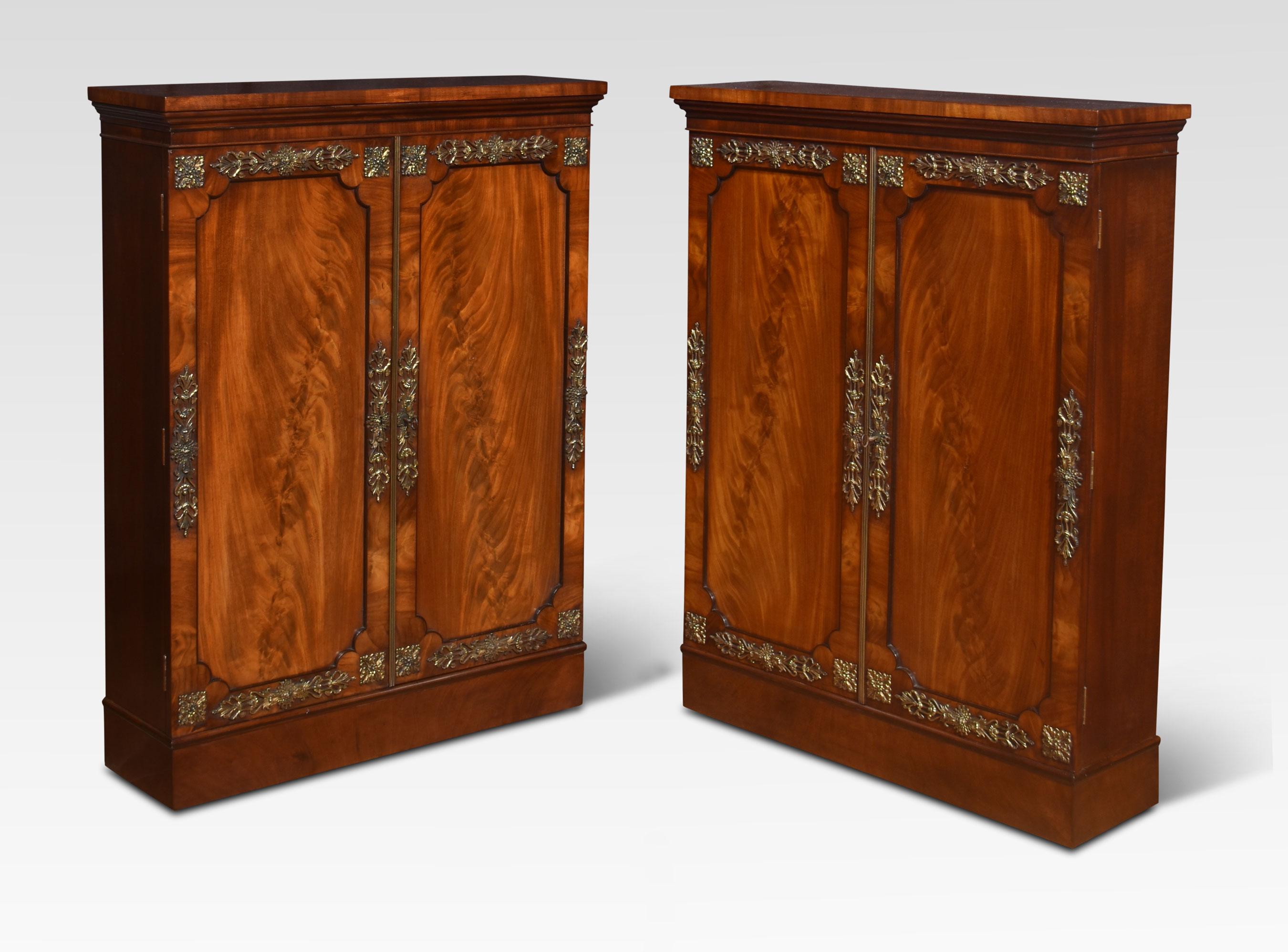 Pair of figured mahogany cabinets, the rectangular tops above a pair of well-figured mahogany doors opening to reveal an adjustable shelved interior. The cabinets are adorned with floral brass mounts and having Brahma locks. All raised up on a