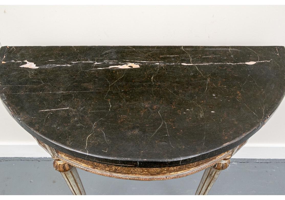 The neoclassical style demi-lune tables with faux black and brown marble painted tops. The carved and gilt aprons with running bands of circles with center rosettes. With a gilt leafy upper framing band, a beaded lower one, and antique pale green