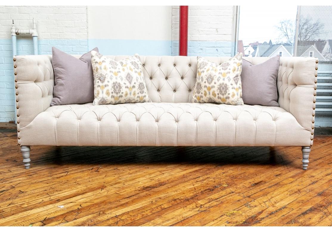 Finely crafted in contemporary lines with square frames and high arms. The backs, inside arms, and seats button tufted in off-white linen like upholstery. The arms with large brass nail head trim. Raised on gray painted turned front legs and square