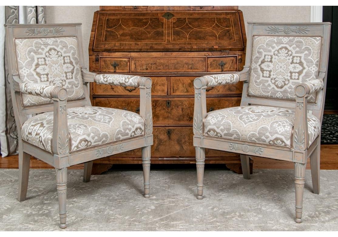 A pair of custom upholstered arm chairs with intentionally distressed paint decorated frames in bluish taupe. The carved palmettes on the arm supports echo the ones on the crest rail. With round armrests with manchettes, web construction with firm