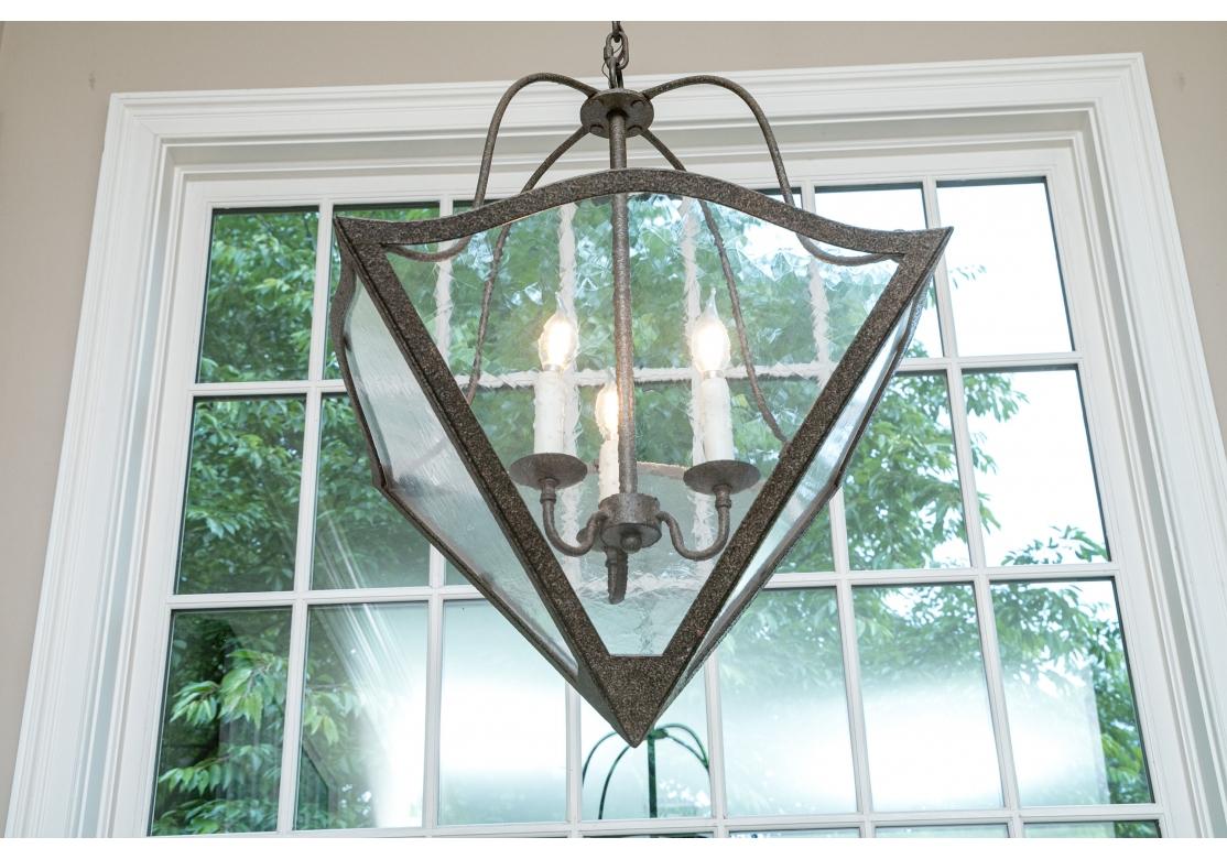 Pair of fine quality hammered or textured iron lanterns with pyramid form having 3 illuminating wax candles.
The lanterns comprised of four scrolling rods conjoined with a serpentine frame holding four panels of textured glass. Come with chain and