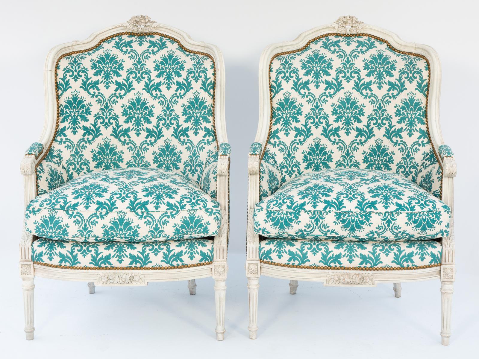 Coppia di sedie bergere dipinte, ognuna delle quali ha una struttura incanalata con finitura verniciata color avorio che mostra un'usura naturale, con la cresta arrotondata sormontata da un intricato getto floreale intagliato, che si arrotonda in