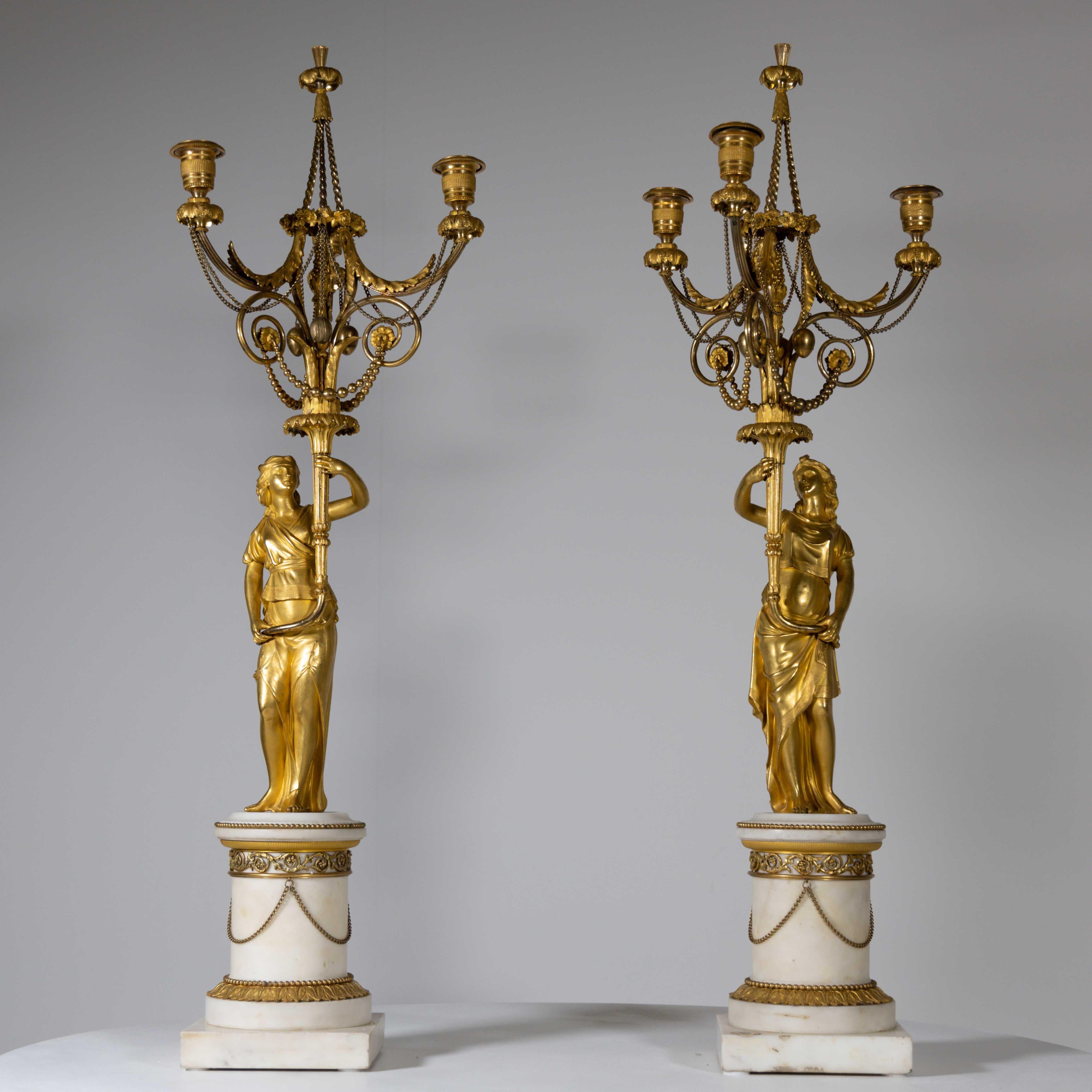 Pair of fire gilded girandoles with female torchbearers on white marble pedestals with gilded bronze applications. The female figures carry the three-flame candelabrum with garlands of link and pearl chains as well as leaf decorations and a central